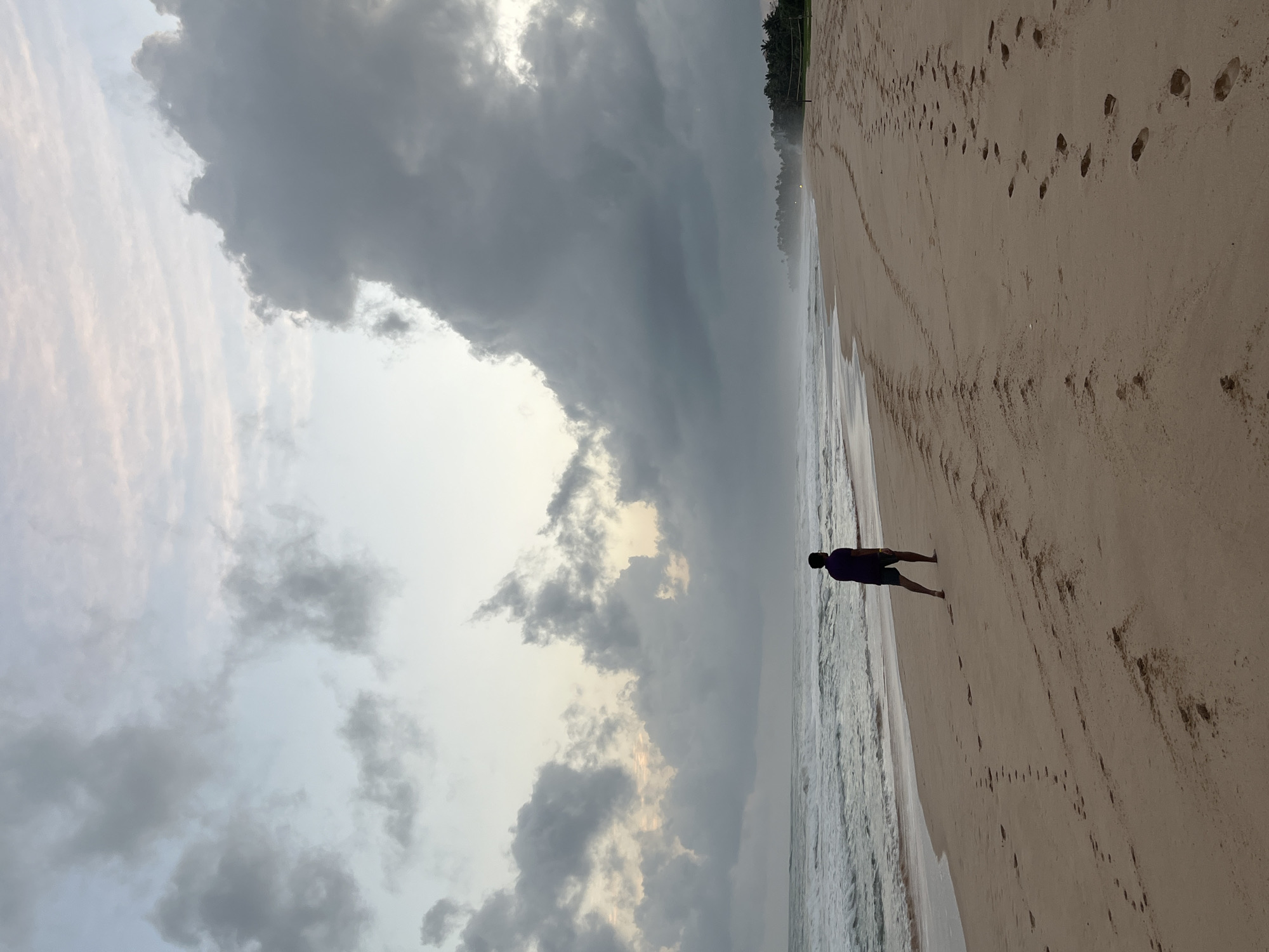 A lone individual, viewed from behind, walks along a sandy beach in Habaraduwa, Sri Lanka. The person is wearing a dark purple t-shirt and dark shorts. The sky is overcast with dramatic, dark clouds that obscure the sun, creating a diffused light. Waves from the ocean gently lap against the shore, and the sand is marked with numerous footprints, indicating previous activity. In the distance, a line of palm trees suggests the presence of vegetation along the coastline. The overall atmosphere is serene and somewhat somber, characteristic of an overcast day by the sea.
