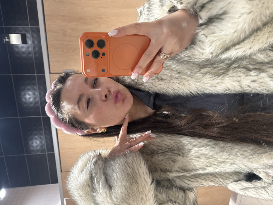 A female individual is depicted in a mirror selfie taken indoors. She has long, dark brown hair and wears a light pink, padded, segmented headband. Her attire includes a light grey/beige faux fur coat worn over a dark grey zippered top. She displays light-colored manicured fingernails, possibly white or light pink, with a glitter effect on the tips. Gold-toned spherical stud earrings are visible. She poses with puckered lips in a "duck face" expression and her right eye (viewer's left) partially closed. Her left hand (viewer's right) is positioned with the index finger touching her jawline. She holds an orange-cased smartphone, equipped with a multi-lens camera array and a circular magnetic attachment on its back, in her right hand (viewer's left). Rings are present on the ring fingers of both hands. The background consists of dark grey/black tiled walls with a faint circular pattern, a silver-toned wall-mounted dispenser (likely a toilet paper or soap dispenser) on the upper left, and a lighter brown, possibly wood-paneled, wall section below the tiles. This environment suggests a public restroom or changing room. The precise location within the city of Akseno-Butyrki, Russia, cannot be determined from the image.