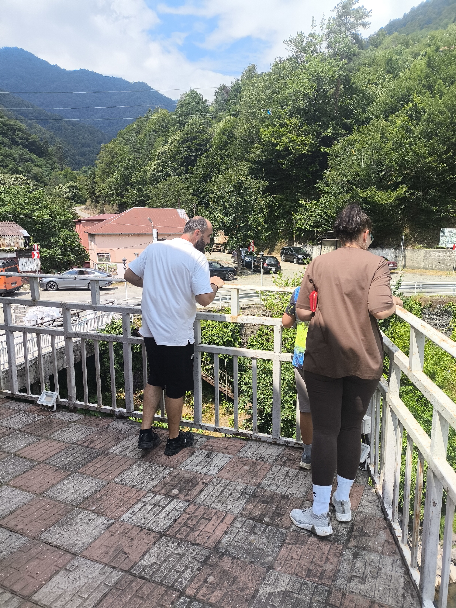 In Zvare, Georgia, on a sunny day, three individuals stand on a tiled pedestrian bridge overlooking a lush, mountainous landscape. A man with a bald head and a white t-shirt and black shorts faces away from the camera, leaning on the bridge's railing. To his right, a woman in a brown t-shirt and dark leggings also looks out at the scenery. Partially visible behind them is a third person wearing a multi-colored shirt. The bridge has a white metal railing and a patterned tile floor. In the background, a winding road is visible, with cars parked and driving. Buildings with red roofs and green trees populate the valley, leading up to a vast, densely forested mountain range under a bright blue sky with scattered clouds.