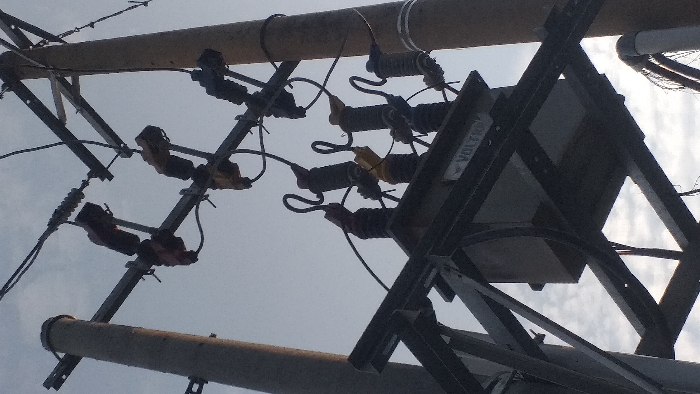 A low-angle, forensic view depicts an electrical power distribution setup in Lamreung, Indonesia. The scene is dominated by two utility poles supporting a suspended electrical transformer. The left pole appears concrete and dark grey, while the right pole is also a utility pole, slightly lighter in tone. A large, rectangular, dark grey electrical transformer, marked with "VOLTRON" on a label, is centrally mounted on a robust black metal framework positioned between the two poles. Overhead metal cross-arms extend from both poles, supporting multiple black, yellow, and brown ceramic or polymer insulators and associated high-voltage fuses or disconnect switches. Numerous black electrical wires connect these components, leading into and out of the transformer. The background consists of a predominantly overcast or cloudy sky, with some lighter patches indicating partial cloud cover. No individuals or active movements are observed.