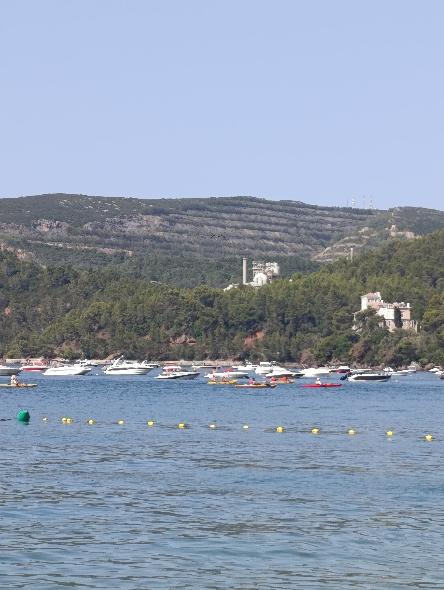 A bright, sunny daytime, full-shot image captures a coastal scene with a wide expanse of blue water in the foreground. A line of yellow buoys stretches across the mid-ground, with a green buoy floating near the left edge. Beyond the buoys, a collection of white and red motorboats are moored in the water. In the background, a densely wooded hill slopes upwards, dotted with a few buildings. A large white industrial complex with smokestacks is visible on the upper left slope of the hill, and a multi-story white building with a red roof is situated on the right side of the hill. The top of the hill is covered in terraced vegetation, and several communication towers are present on the crest. The sky is clear and light blue. The location is the coast of Setúbal, Portugal, likely near the Arrábida Natural Park.