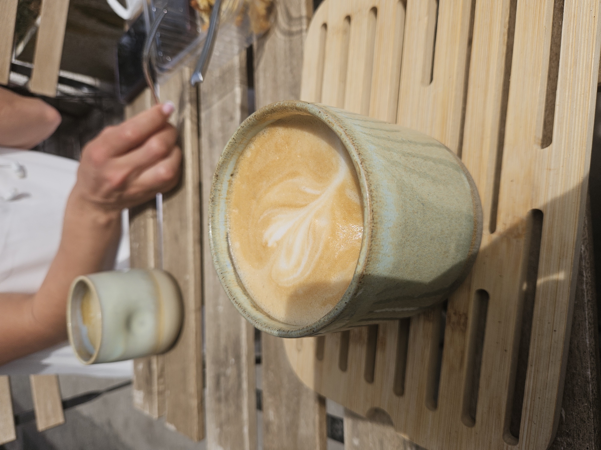 The image presents a rotated overhead view of an outdoor cafe table, focused on two coffee cups and a person's hands. The central subject is a textured, light green ceramic coffee cup filled with a latte, prominently displaying a white heart-shaped latte art design on its frothy surface. This cup is placed on a light-colored wooden slatted placemat or tray.
To the left of the main cup, a person's forearm and hand are visible, holding a slender metal utensil, possibly a spoon or fork. The hand has neatly manicured nails with light pink polish. Further back on the table, another, smaller ceramic cup with a similar design is partially visible and out of focus. In the background, a blurry plastic container, potentially holding food, can be seen, along with another person's arm.
The setting suggests an outdoor environment, possibly a cafe or patio, characterized by natural sunlight casting subtle shadows across the wooden table and slatted surfaces. The bright lighting indicates it is daytime, likely morning or early afternoon. The overall atmosphere appears relaxed and casual, centered around enjoying coffee and a potential meal. There is no visible text in the image.