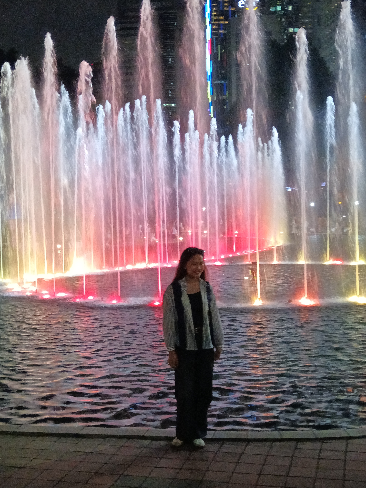 A woman stands in front of a brightly lit musical fountain in Kuala Lumpur, Malaysia. The fountain displays multiple jets of water illuminated with vibrant red and yellow lights, creating a dynamic and colorful spectacle. The water surface reflects the lights, adding to the visual appeal. Behind the fountain, tall buildings with illuminated windows are visible, indicating an urban setting at night. The woman is wearing a dark top, a striped cardigan, and dark pants. She is looking towards the viewer with a slight smile. The foreground shows a tiled pathway and the edge of the fountain basin. The overall scene suggests a lively evening in a city center.