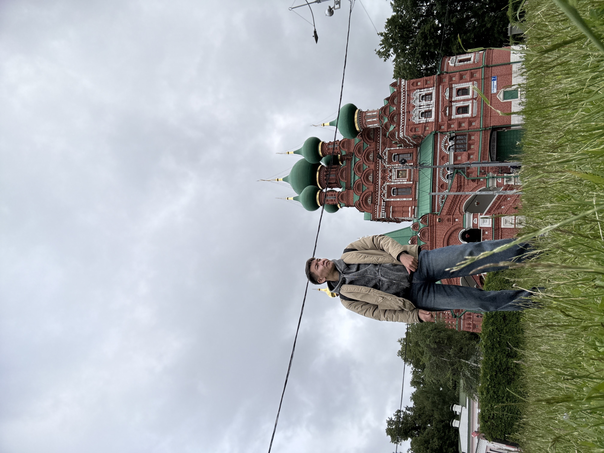 A man in a beige jacket and blue jeans stands on the ground, looking up at the Church of the Intercession of the Holy Virgin in Khimki, Russia. The red brick church with green domes stands behind him, partially obscured by tall grass in the foreground. The sky is overcast. Power lines cut across the scene.