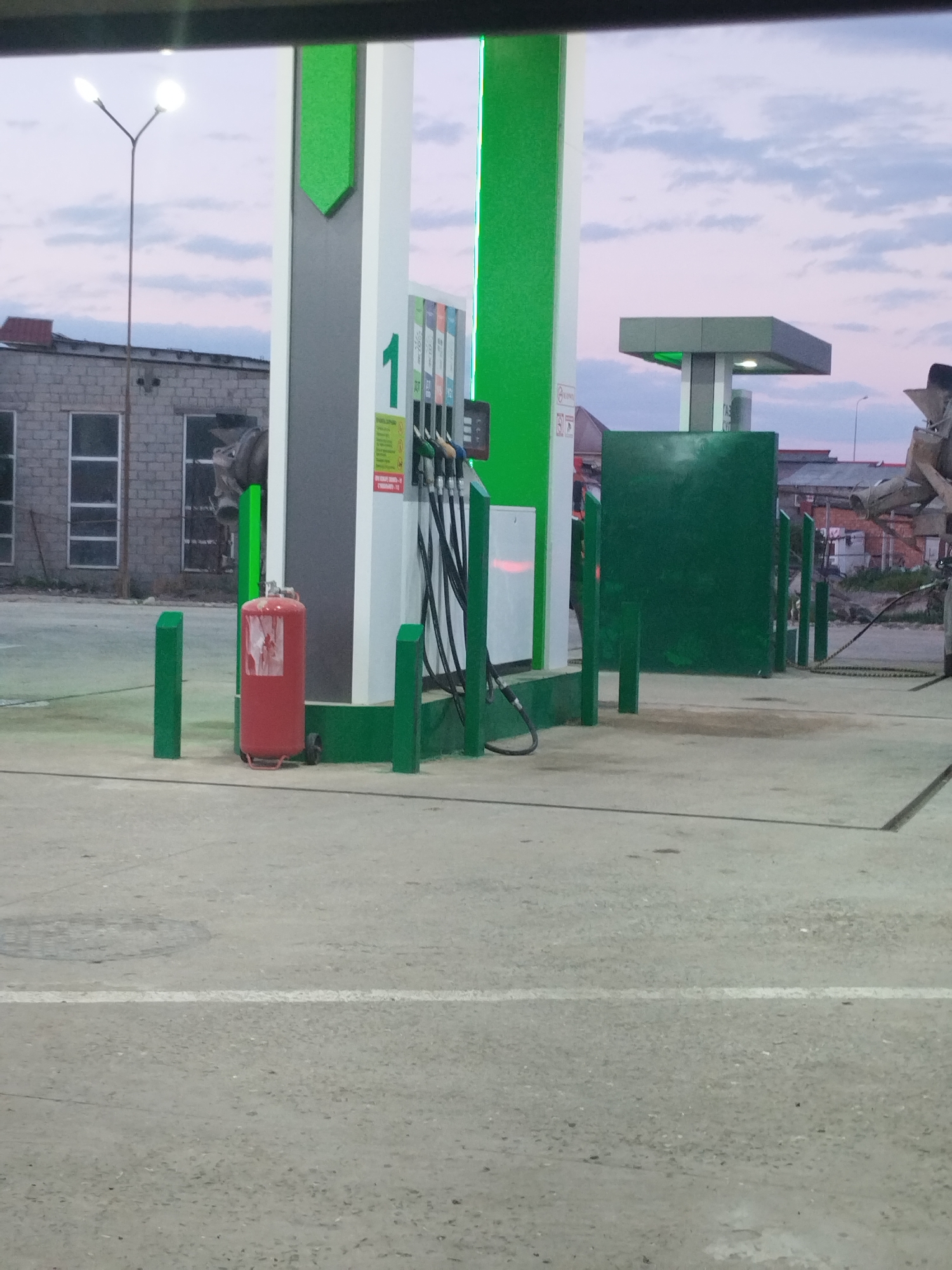 The image depicts a gas station in Kala, Russia, during twilight. The sky is a soft lavender and pink, with scattered clouds, indicating either dawn or dusk. The primary subject is a modern gas pump station with white and bright green accents. The pump itself has multiple hoses and nozzles. To the left of the pump, a red fire extinguisher stands prominently on the concrete forecourt, surrounded by short, green bollards.
In the background, a utilitarian brick building with several large, dark windows is visible. To the right of the pump, a large, dark green panel stands, also bordered by green bollards. Above this, a minimalist canopy structure with grey and green elements is perched on a central pillar. Further in the background, faint outlines of other buildings and a lamppost can be seen against the dusky sky. The overall scene suggests a quiet, functional environment. There is some faded text on the fire extinguisher and on signage on the pump, but it is not legible. The lighting suggests a low-light condition, characteristic of twilight.