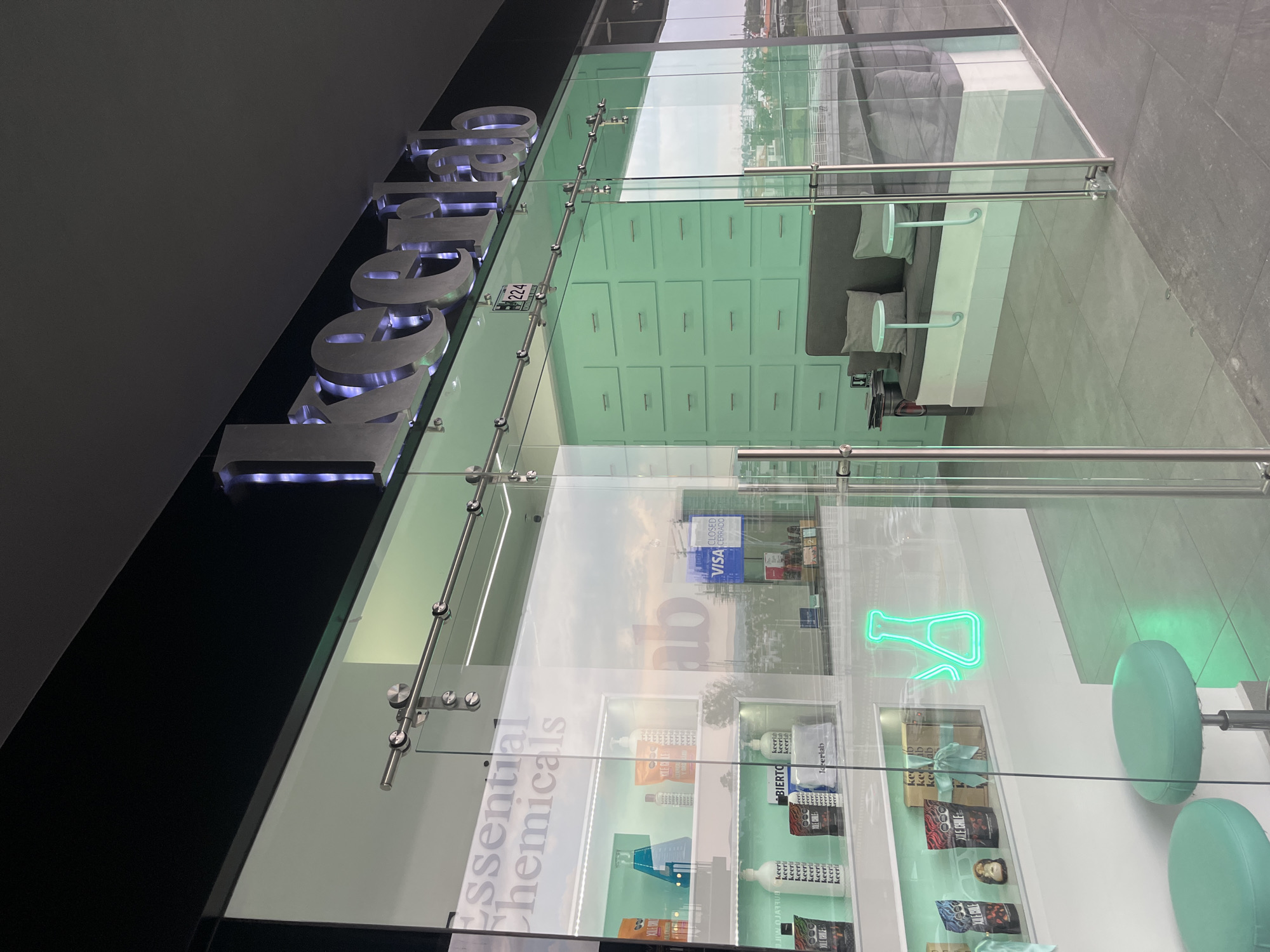 This media file captures an angled view of the exterior and interior of a modern commercial establishment, likely a shop, through a large glass facade. The setting is San Bernardino Tlaxcalancingo, Mexico.
The main subject is the storefront, characterized by a large sign with illuminated silver block letters spelling "keerlab" mounted above the entrance. The entrance itself is a glass sliding door system with polished metal fittings. A small white sign bearing the number "224" and a wheelchair accessibility symbol is attached to the glass.
Inside the store, a contemporary and minimalist aesthetic prevails, featuring a dominant light green and white color scheme. To the left, a wall is adorned with numerous light green, drawer-like panels. Adjacent to this, a seating area includes a grey sofa with throw pillows and two small, round white tables, suggesting a waiting or lounge space. On the right, through the transparent glass, display shelves are visible, stocked with various products. Many items are labeled with "keerlab," and other visible texts include "Essential Chemicals," "keeri," "YILE CHILE," and "000." A striking green neon sign in the shape of a chemical flask or beaker adds a distinctive touch. Two light green, round stools are also seen near the display areas.
The glass facade causes significant reflections of the exterior environment, including a bright sky with clouds, what appears to be an outdoor area like a street or parking lot, and other structures. Visible reflected text includes "VISA" and "CLOSED CERRADO," possibly indicating the store was not operating at the time of the photo or reflecting signage from nearby businesses. Another reflection shows the letters "ab." The scene is static, with no people present, suggesting a daytime shot of the store's architecture and interior design.