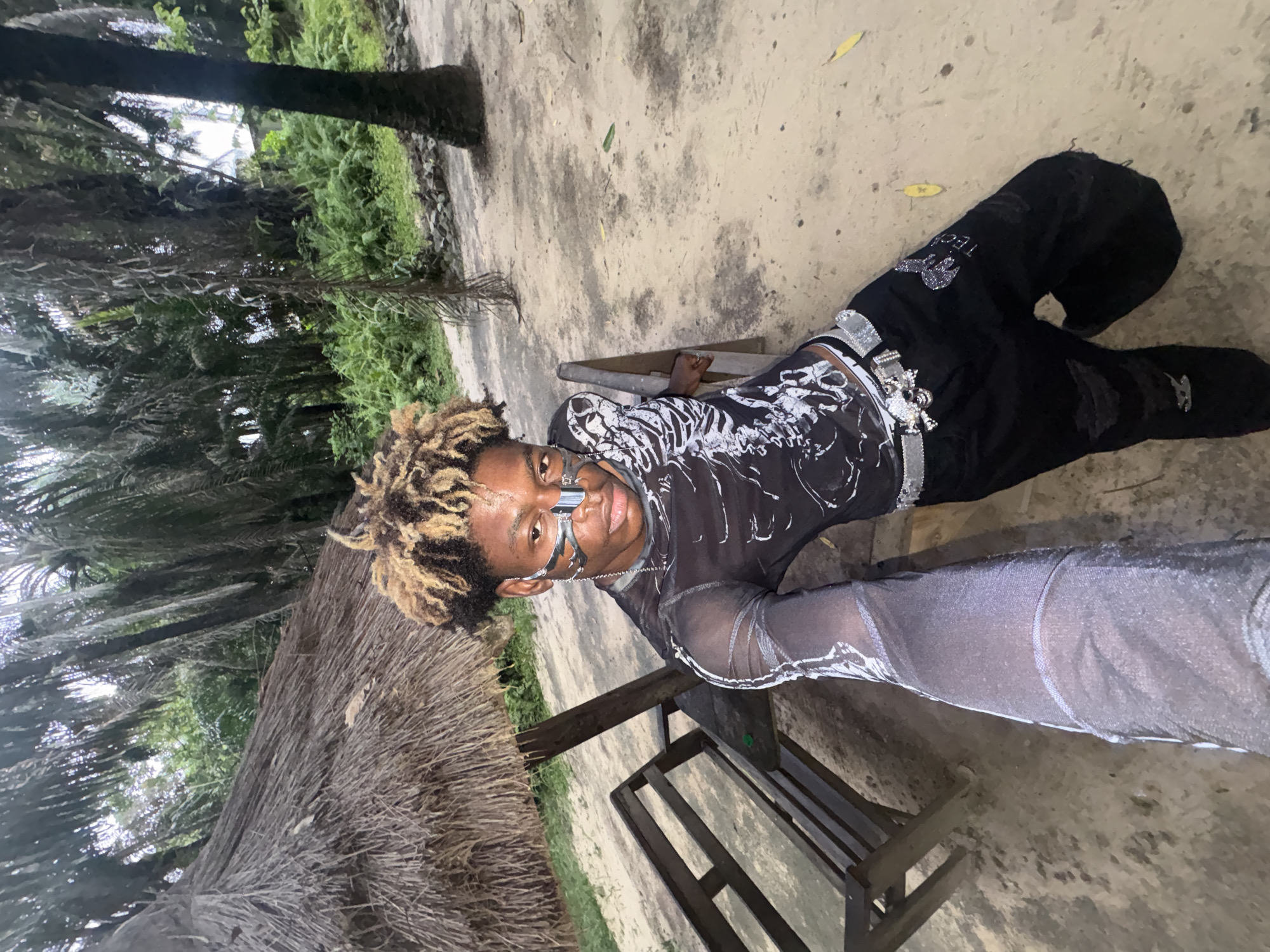 A person with bleached blond hair and black clothing is lying on a wooden bench outdoors. They are wearing a sheer top with a white skeleton print, black pants with silver embellishments, and a silver mask over their nose and eyes. The person is facing the camera. The bench is situated in a sandy area with vegetation, including palm trees and shrubbery. The photo was taken outdoors, likely in Sangotedo, Nigeria.