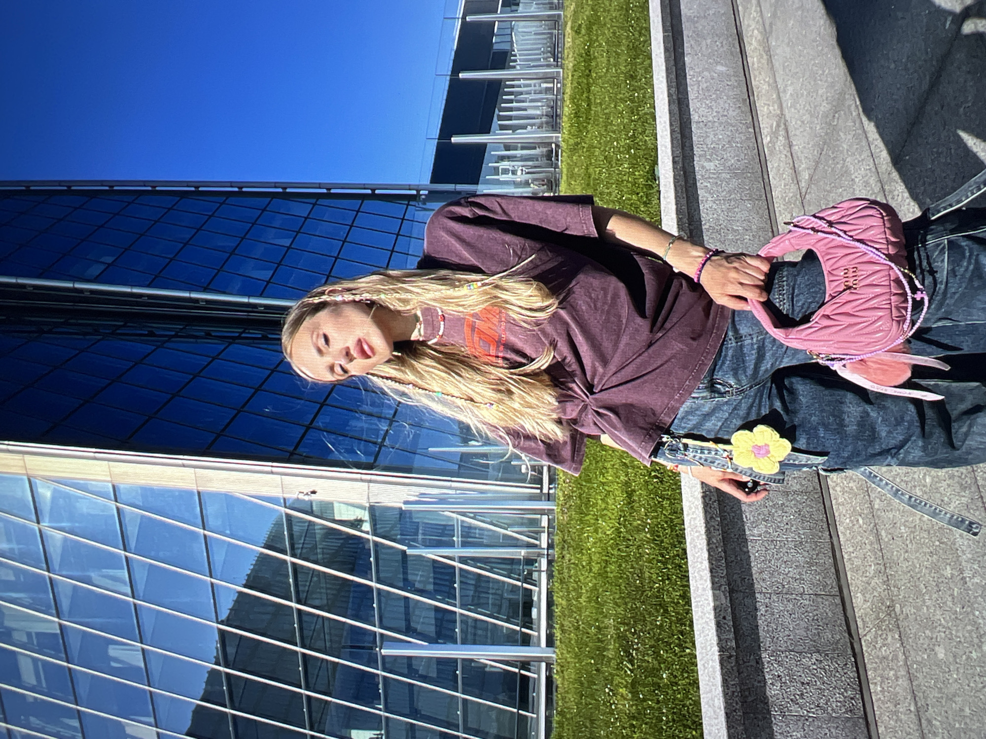 A young woman with blonde hair, styled in braids with colorful beads, poses in front of a modern glass building. She is wearing a dark purple oversized t-shirt, twisted at the front, and loose-fitting blue jeans. The t-shirt has a graphic print on it. She is accessorized with a beaded necklace, bracelets, and is holding a small, pink quilted handbag. A yellow flower keychain is attached to her jeans. The background consists of a lush green lawn and a large, angular glass building reflecting the bright blue sky. The scene appears to be an outdoor urban environment on a sunny day. The location is identified as Unknown, Russia.