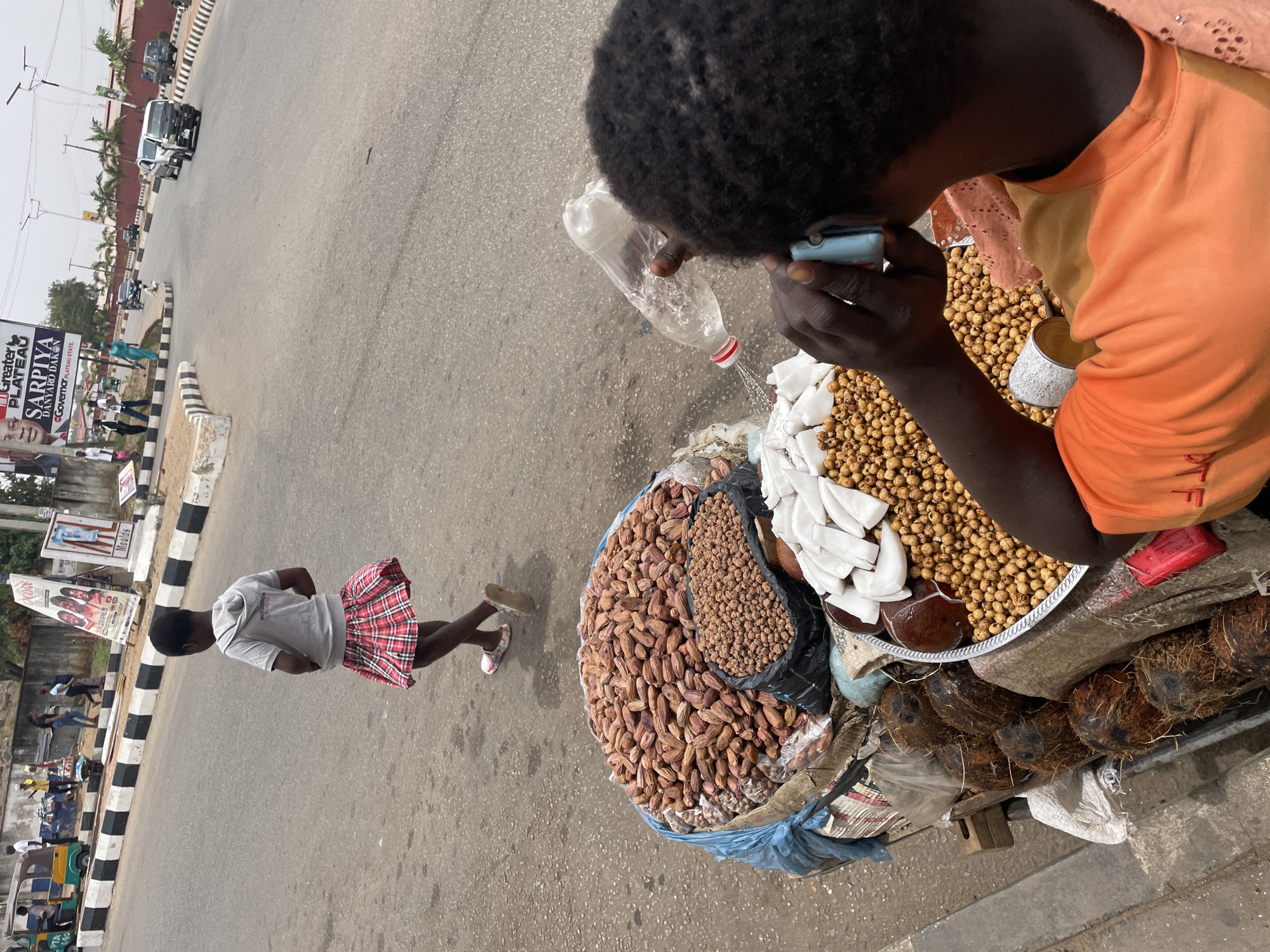 An overhead, slightly tilted photograph captures a vibrant street scene, likely a market or roadside vending area in Jos, Nigeria, during the daytime.
The primary subject is a vendor, seen from behind and the side, with dark, curly hair and an orange t-shirt. The vendor is leaning over a large, round tray filled with various goods. In their hands, they hold a clear plastic water bottle from which water is being poured, and a small blue object, possibly a mobile phone or a small container, held to their ear or mouth.
The tray displays a substantial amount of brown, elongated dates, a smaller pile of spherical, light brown tiger nuts, and white, sliced pieces that appear to be coconut meat. Below the tray, several whole coconuts are stacked. A small, red rectangular object is also visible on the tray.
Further into the scene, a young girl, dressed in a grey t-shirt and a red and white plaid skirt, walks away from the vendor on the paved road. The road features black and white striped curbs. In the background, other vehicles, including cars and a commercial tricycle ("keke napep"), can be seen, indicating a moderately busy thoroughfare. Palm trees and buildings line the street in the distance.
On the left side of the road, several billboards are visible. One prominent billboard displays political campaign information, including the text "GREATER PLATEAU SARPIYA DAKUR & DAKUR" and "Governor PLATEAU STATE," suggesting an ongoing election campaign. The lighting is bright and even, indicating clear weather.