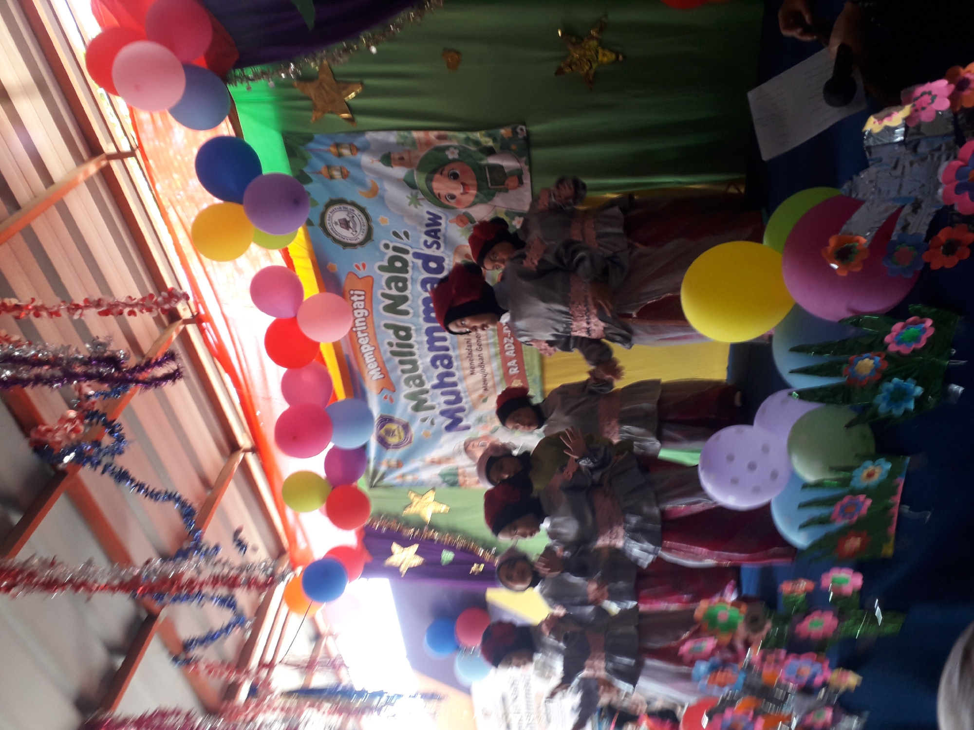 This is a medium shot, eye-level, outdoor, daytime photograph of a group of children performing on a stage. The stage is decorated with colorful balloons, tinsel streamers, and a banner that reads "Memperingati Maulid Nabi Muhammad SAW". The children are wearing traditional Islamic clothing. They are standing in front of a green curtain and a banner. There are also some decorations on the table in front of the stage, including flowers and balloons. The image was taken in Cankuang, Indonesia.