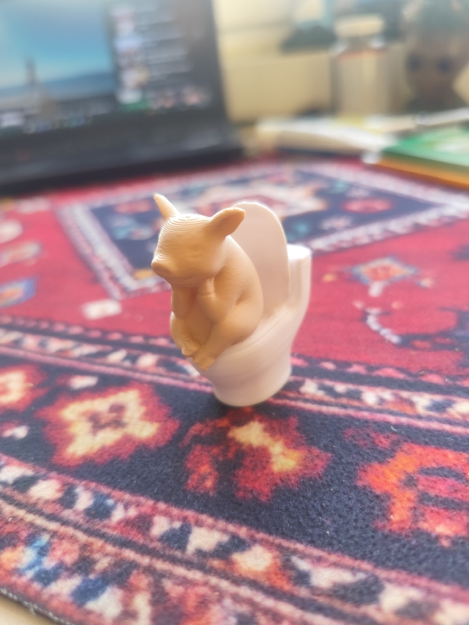A 3D-printed pig figurine is sitting on a white toilet seat, resting on a patterned rug. A laptop and other objects are blurred in the background. Location cannot be inferred.