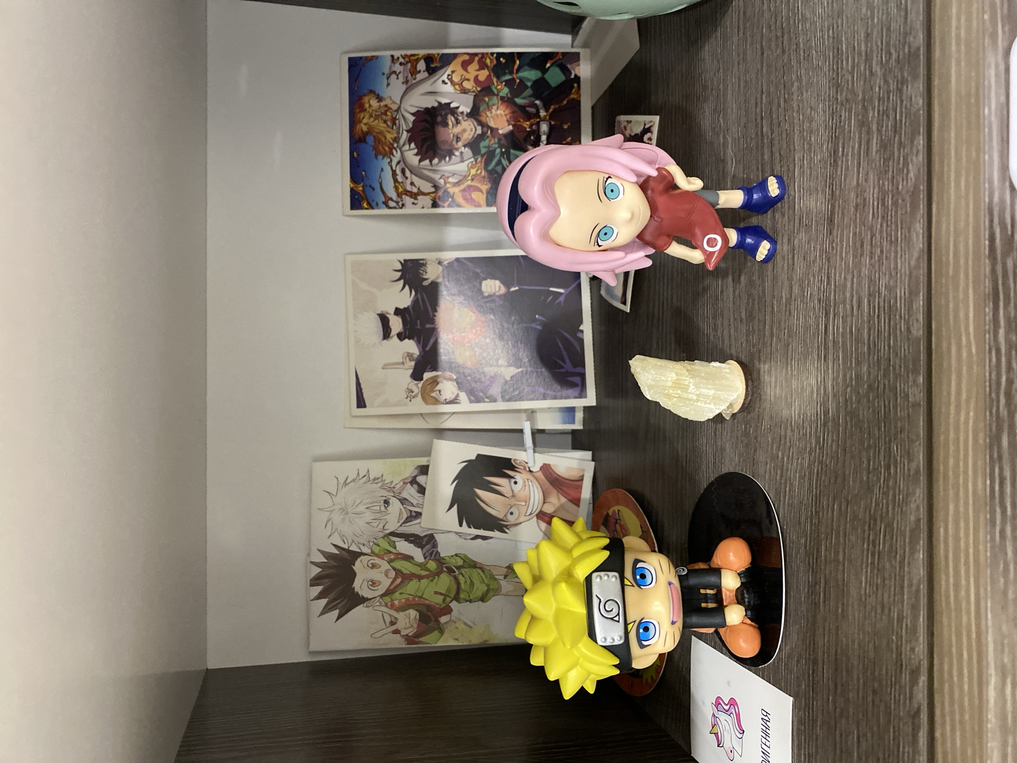 The image shows a shelf containing anime-themed items. From top to bottom: a card featuring characters from Demon Slayer, a Sakura figurine, a card featuring characters from Jujutsu Kaisen, a piece of bone or seashell, a card featuring Luffy from One Piece, a card featuring characters from Hunter x Hunter, and a Naruto figurine. The background suggests the shelf is part of a built-in unit, likely located in an interior space. There is no indication of a specific city or people.