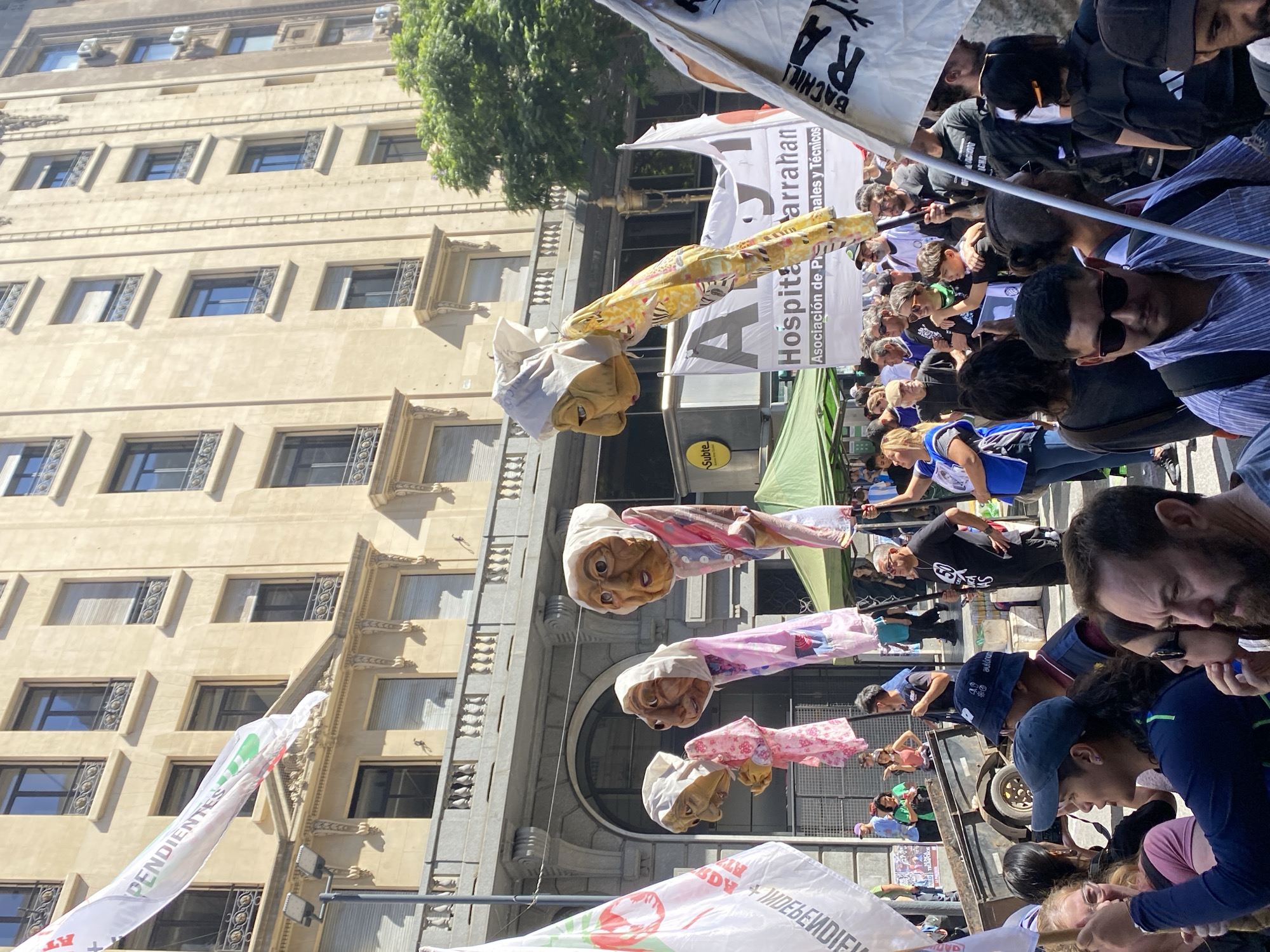 The image depicts a crowded public gathering or protest taking place on a sunny day in downtown Buenos Aires, Argentina. The background features several tall, light-colored, multi-story buildings displaying classical European architectural elements, including numerous rectangular windows, some with closed external blinds, and ornate wrought-iron balconies. Green tree foliage is visible on the right side.
The foreground and midground are densely populated by a diverse crowd of individuals, varying in age and gender, dressed in casual clothing. Many individuals are holding poles, flags, or banners. Three prominent, large puppets, crafted to resemble elderly women with detailed, wrinkled faces and white head coverings, are elevated vertically above the crowd. These puppets are dressed in patterned fabrics, specifically pink floral, purple patterned, and yellow patterned garments.
Key visible signage includes:
* A large white banner in the midground reads "Hospital Garrahan" with "Asociación de Profesionales y Técnicos" printed below it, indicating the subject of the gathering.
* A yellow sign displaying "Subte" in blue text is located beneath the "Hospital Garrahan" banner, signaling a subway entrance.
* A partially visible white banner in the upper left section features the word "PENDIENTES" in red and green text.
* Multiple white flags are dispersed throughout the crowd; one in the lower right foreground shows "BACHIL..." and "RA" in black print, while another in the left foreground displays red and green circular designs with text including "INDEPENDIENTE."
Other objects include a green canopy or tent structure behind the main banners on the left, and a dark-colored utility trailer with a single visible wheel, also on the left. The crowd appears engaged in the protest, with individuals standing, conversing, and observing, under direct sunlight.