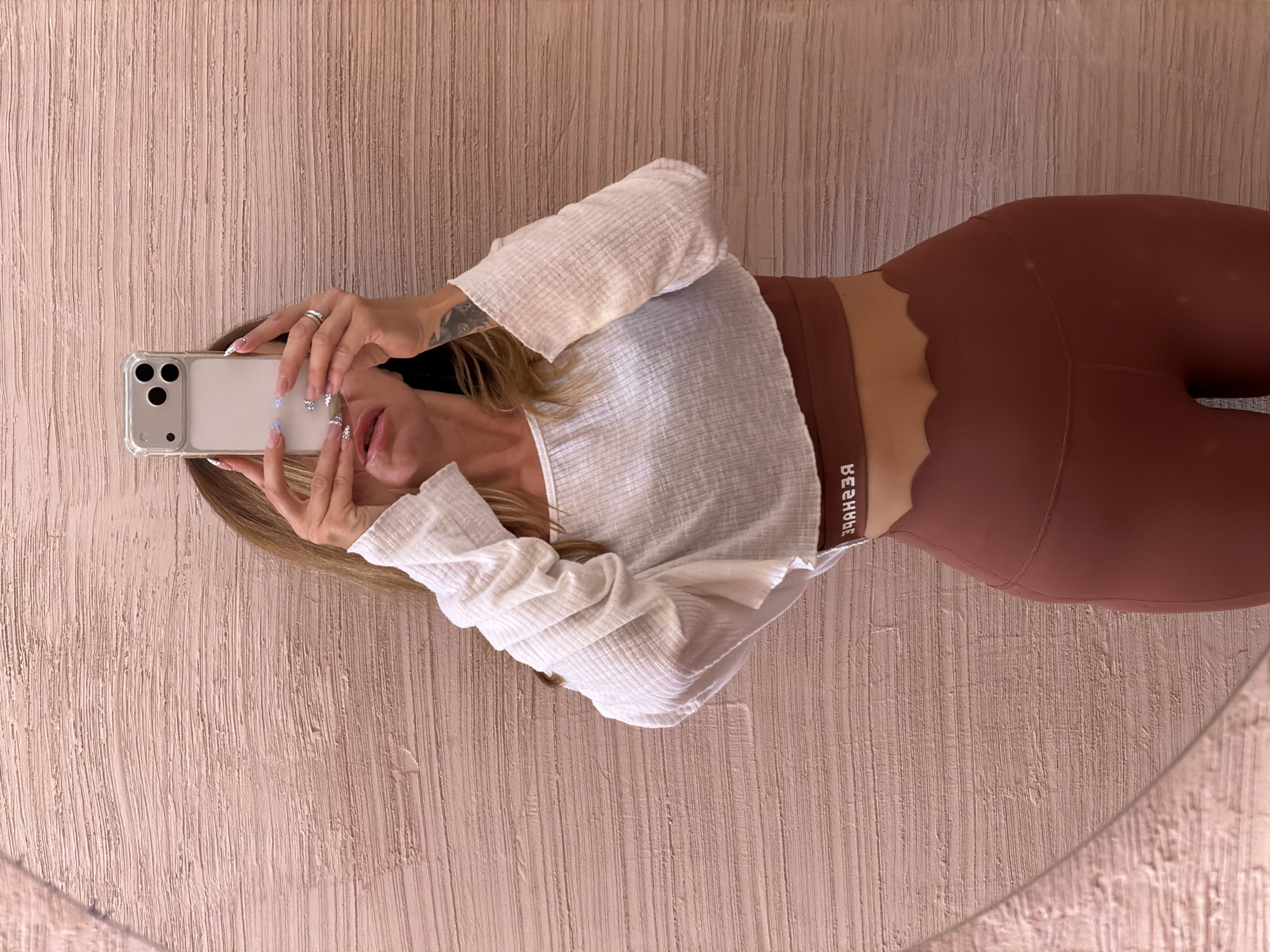 A female adult is taking a selfie in front of a mirror. She is wearing brown leggings with a scalloped waistband and a white, long-sleeved shirt. She is holding a smartphone in front of her face. Her hair is blonde. The background consists of a textured, light pink wall. Based on the text on her clothing, "BEREHYBE," it's possible she's in a location associated with that brand. There is no definitive evidence that the photo was taken in North Kuta, Indonesia.