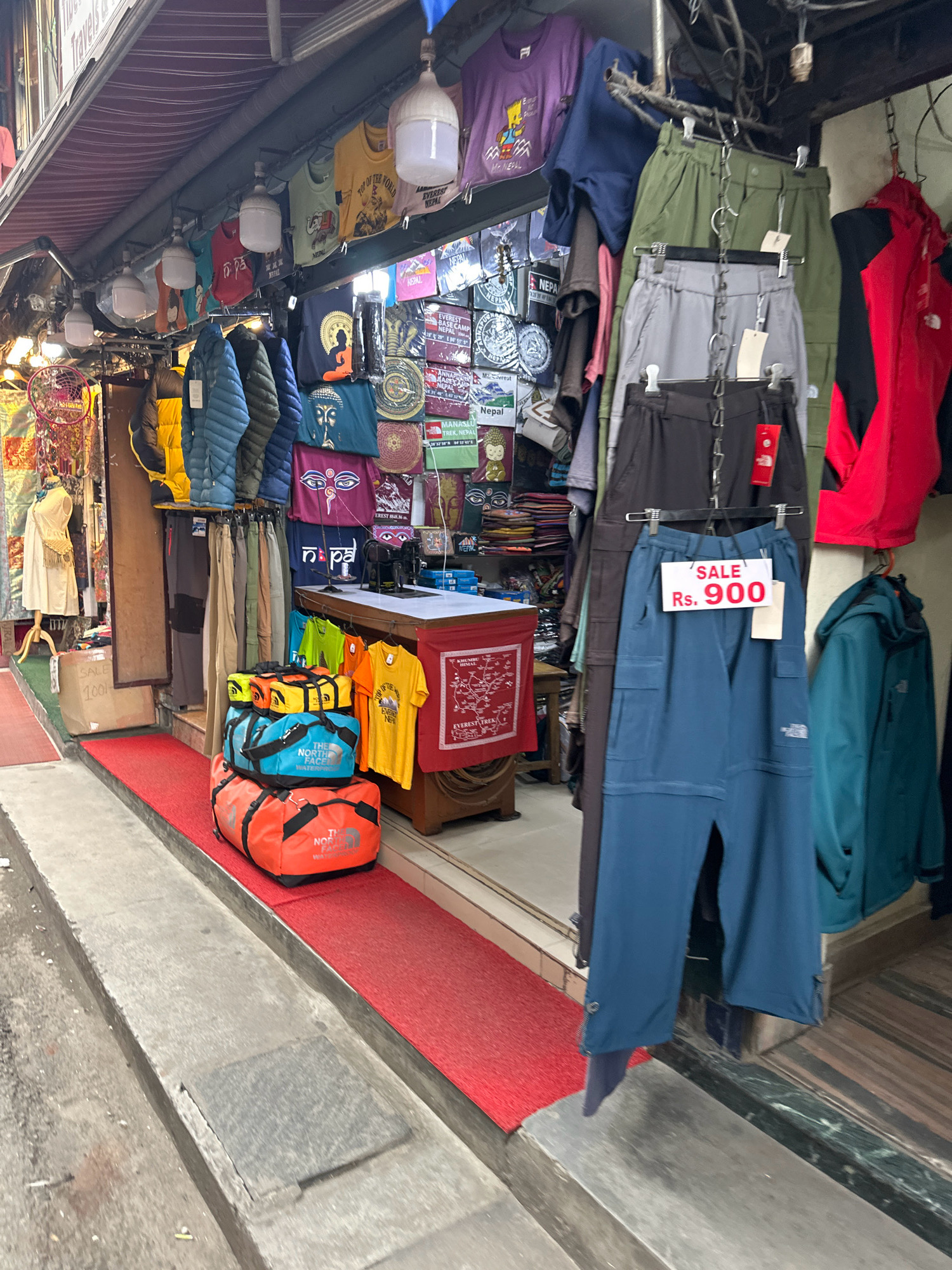This is a street scene in Kathmandu Metropolitan City, Nepal, showcasing a retail establishment with an awning. The store displays a variety of merchandise, including apparel such as t-shirts, jackets, and pants, as well as luggage. Some items are hung from racks, while others are stacked. A mannequin is visible near the entrance. The items for sale appear to be aimed at tourists, with many featuring images and text related to Nepal and Mount Everest. A sign with "SALE Rs. 900" is prominently displayed on a pair of blue pants. The ground in front of the store is covered with a red carpet. The overall impression is that of a bustling market stall catering to visitors.