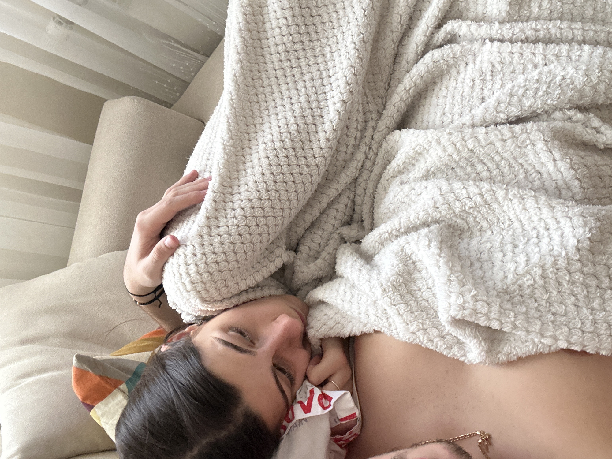 A person lies under a large white, textured blanket on a beige couch with a colorful patterned pillow. The person's face is visible, with dark hair, closed eyes, and a white cloth with red writing near their mouth. The person's bare shoulder and a gold necklace are also visible. The location appears to be indoors with natural light coming through a window with white curtains. There is no information to suggest this image was taken in Plovdiv, Bulgaria.