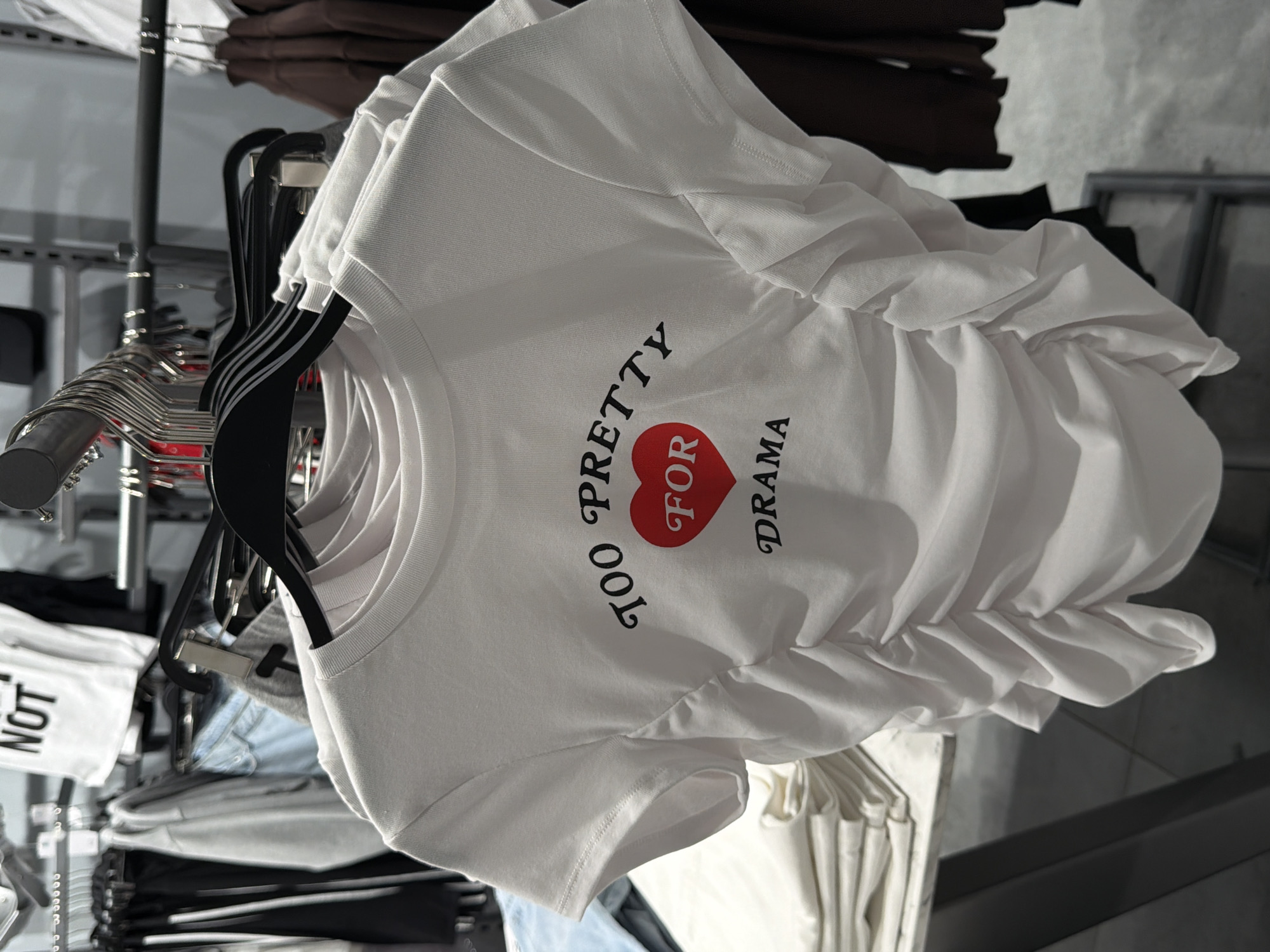 A white ruched t-shirt with the text "TOO PRETTY FOR DRAMA" and a red heart graphic is hanging on a rack in a clothing store. The t-shirt is the primary focus of the image. In the background, other clothing items are visible on hangers, as well as shelving units and clothing racks. The overall environment suggests a retail setting. The image was taken in Moscow, Russia.