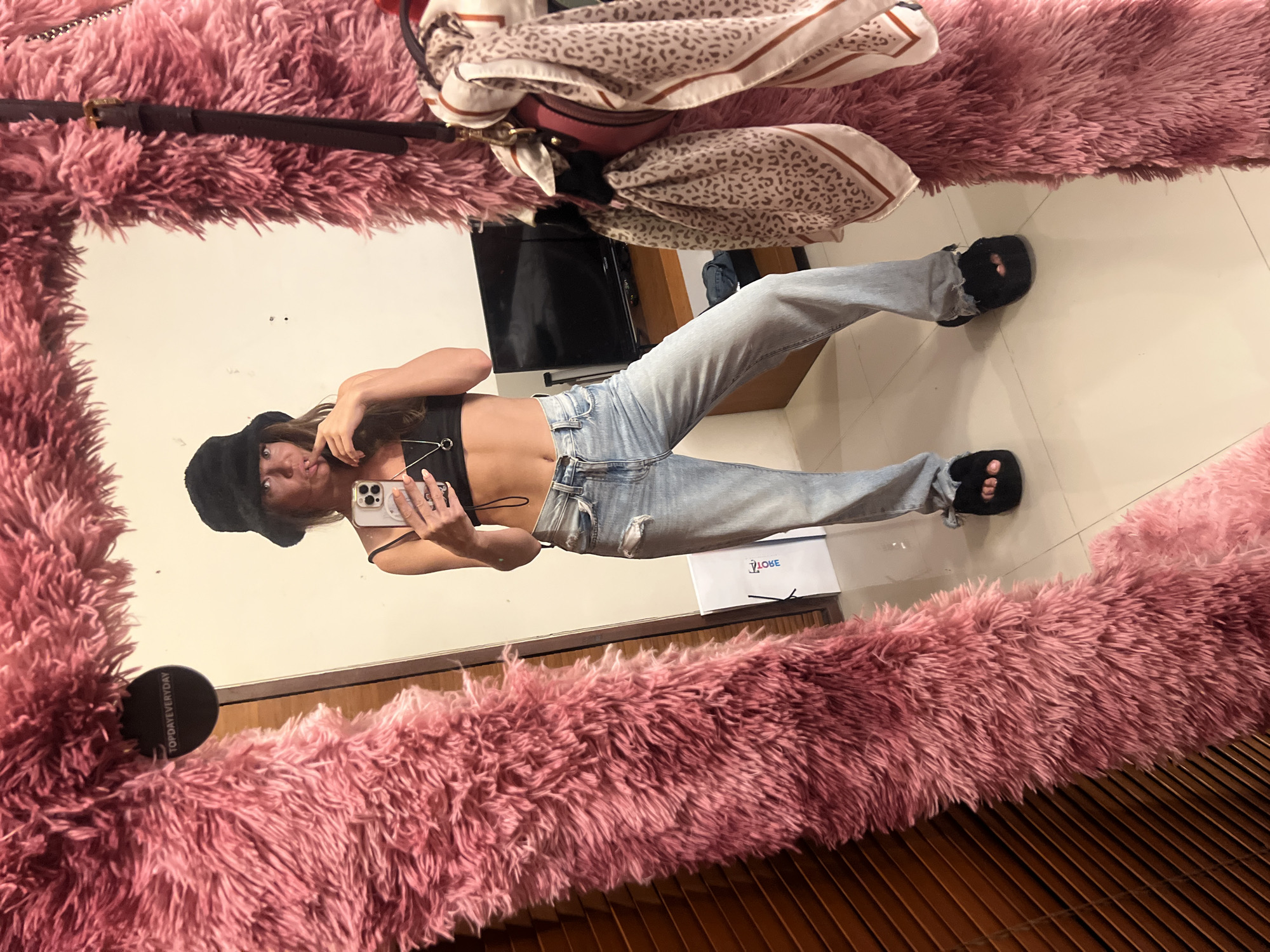 A woman is taking a selfie in a mirror with a fluffy pink frame. She is wearing a black crop top, light blue jeans, and black slippers. She also has a black hat on her head and is holding a smartphone in her right hand. The mirror reflects a room with a TV and a bag on top of the mirror. A "YLOBE" box is on the floor next to the woman. The location appears to be indoors, with tiled flooring and wooden panels. The city of North Kuta, Indonesia, cannot be confirmed from the image.