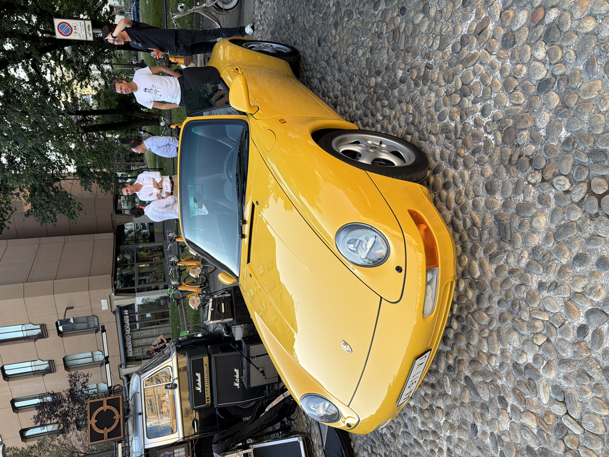 A yellow Porsche 911 is parked on a cobblestone street in Milan, Italy. Several people are gathered around the car, some leaning against it and others standing nearby. The scene appears to be a casual social gathering, possibly related to a car show or a meet-up. The time of day seems to be late afternoon or early evening, with warm, diffused lighting. The weather appears pleasant.
The car is a bright yellow convertible with a black interior. It has a sleek, sporty design with distinctive pop-up headlights. Visible text on a sign near the car reads "0-24" and includes a circular red symbol with a diagonal line through it, indicating a no-parking zone. Another sign on a building in the background reads "ROBERTO ROGARI". In the background, there are trees, buildings, and bicycles parked. Some audio equipment, including what appears to be Marshall amplifiers, is also visible near the car. The overall atmosphere seems relaxed and convivial.