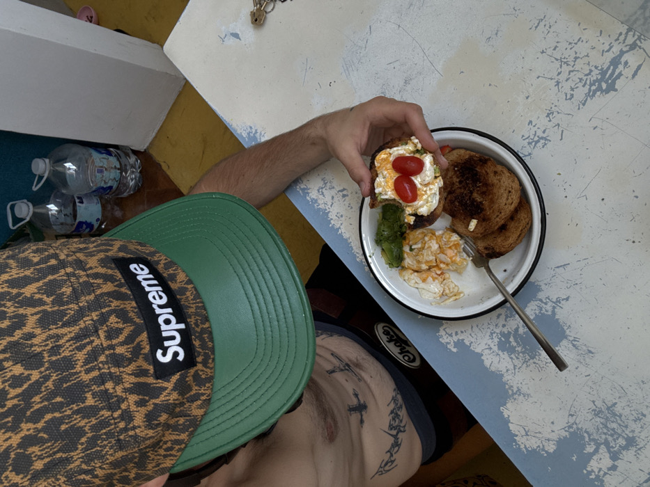 A person wearing a Supreme hat holds a plate with food. The plate contains toast, scrambled eggs, sliced avocado, and a piece of toast topped with a spread and two cherry tomatoes. A fork is on the plate. Two water bottles sit near the table. The person is positioned at a blue and white table. Tattoos are visible on the person's torso. The table has keys and a lock on it.