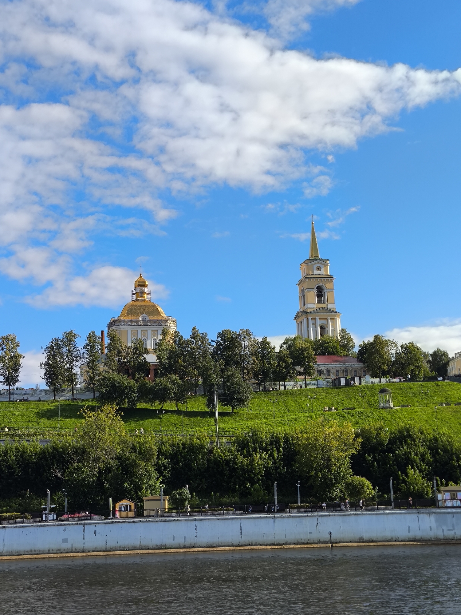 The image shows a wide, calm river in the foreground, with a concrete embankment and a walkway along its edge. Beyond the embankment, a steep, grassy hill rises, topped by a prominent building with a golden dome and a tall bell tower with a golden spire. The buildings appear to be historical religious structures. Trees are scattered across the hillside and around the buildings. The sky is bright blue with scattered white clouds. Several people are visible walking on the embankment and the walkway. The scene is bathed in sunlight, suggesting it is daytime. This is the Spaso-Preobrazhensky Cathedral complex on the embankment of the Kama River in Perm, Russia.