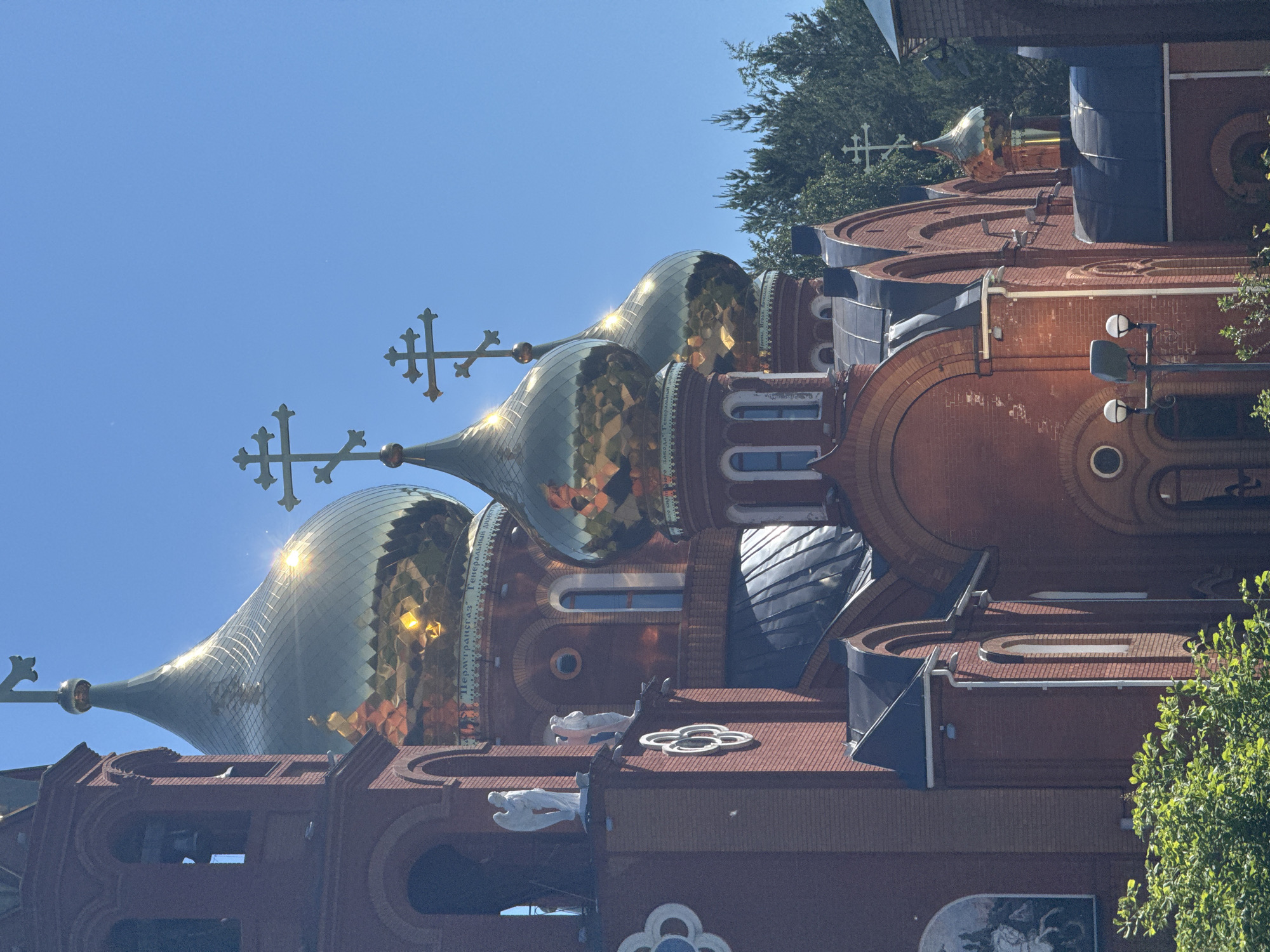 The image depicts a close-up view of the golden domes and red brick architecture of the Holy Trinity Cathedral in Chaikovsky, Russia, under a clear blue sky. The cathedral features multiple onion domes topped with Orthodox crosses, reflecting the bright sunlight. The red brick facade is adorned with arched windows and decorative elements. In the foreground, lush green foliage is visible, suggesting a pleasant outdoor setting. Two white angel statues are positioned on the left side of the cathedral, adding to its spiritual and artistic appeal. The overall scene conveys a sense of grandeur and tranquility, characteristic of religious landmarks.