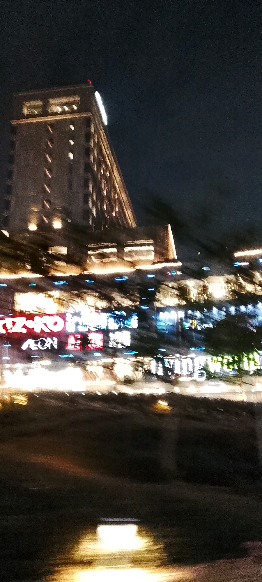The image captures a blurred nighttime view of a building, likely a hotel, in Gunung Putri, Indonesia. The building is tall and illuminated with warm lights, featuring rows of windows and a pointed roof. Below the building, a street level view shows a blur of lights from signage and storefronts. Among the visible signs, "RIZOKO" and "AEON" are discernible, suggesting a commercial area. The overall impression is one of a bustling urban environment at night, with motion blur indicating movement, possibly from a vehicle. The dark sky and the artificial lights dominate the scene, creating a sense of depth and activity.