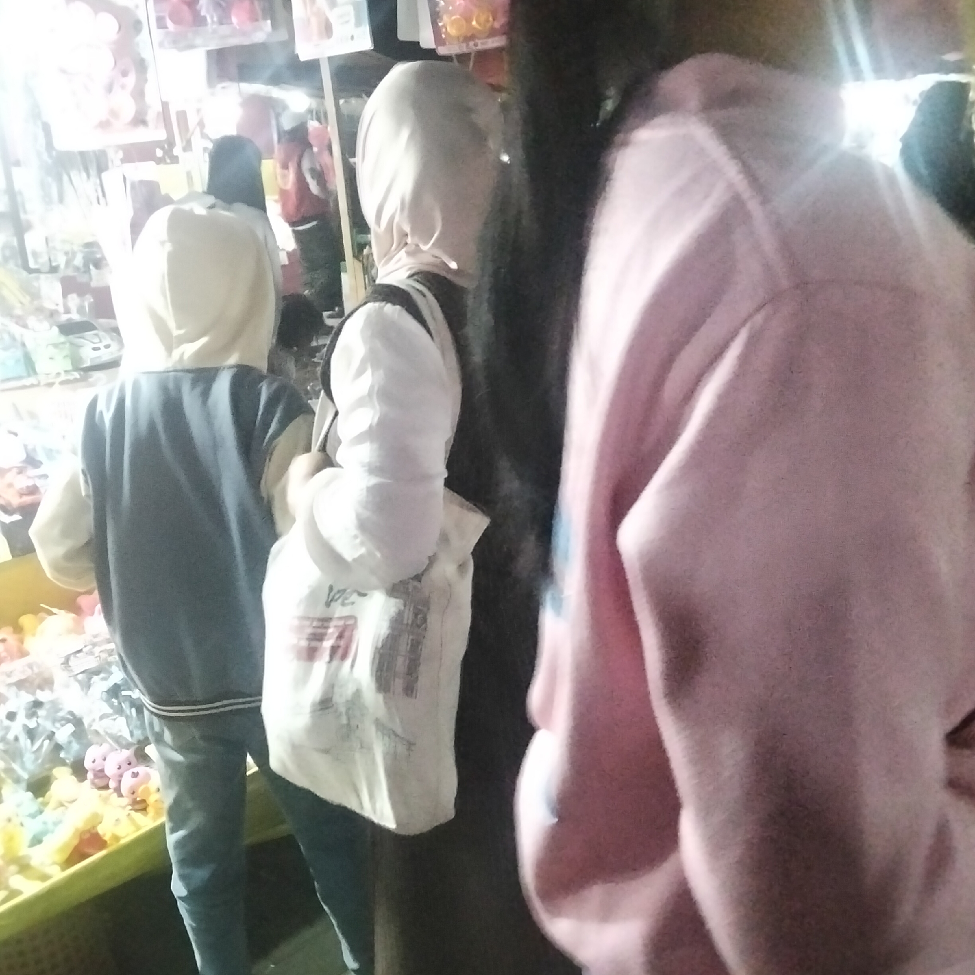 The image shows a street scene at night in Cisaat, Indonesia, possibly near a market or food stall. Three individuals are visible, two in the foreground and one partially obscured in the background. The person on the left is wearing a blue jacket with a light-colored hood and dark pants. They are facing away from the camera, and their hands are not visible. To their right is another person wearing a light-colored hijab and a white shirt, holding a white bag with some writing on it. This person is also facing away from the camera and is partially obscured by a third person on the far right. This third person is wearing a pink hooded sweatshirt and is facing towards the right side of the frame, with their face not visible. The background shows shelves or displays with various items, likely merchandise. The lighting is dim, suggesting it is nighttime, and there are some bright lights illuminating the scene from above and the left. The overall impression is that of a casual gathering or shopping scene in a public area.
