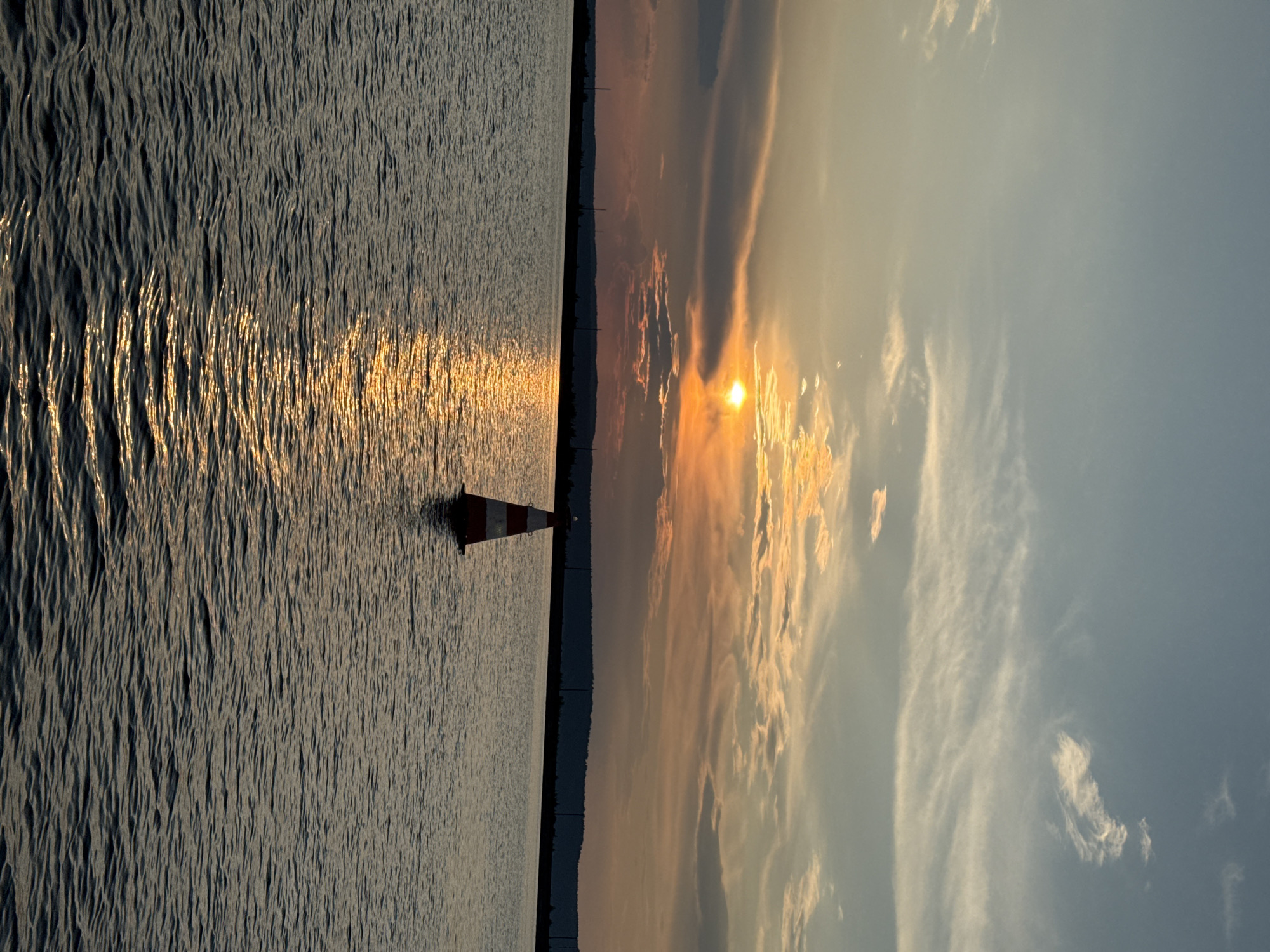 An outdoor, eye-level, medium shot captures a serene sunset over the Kama River in Chaikovsky, Russia. The sun, a brilliant orange orb, is partially obscured by wispy, elongated clouds painted in shades of orange, yellow, and gray. A striped red and white buoy stands alone in the calm, rippling water, its reflection shimmering on the surface. In the distance, a dark, tree-lined horizon stretches across the frame, punctuated by the silhouettes of utility poles. The sky transitions from a pale blue near the top to a warm, sunset-infused palette closer to the horizon. The overall scene evokes a sense of peaceful solitude.