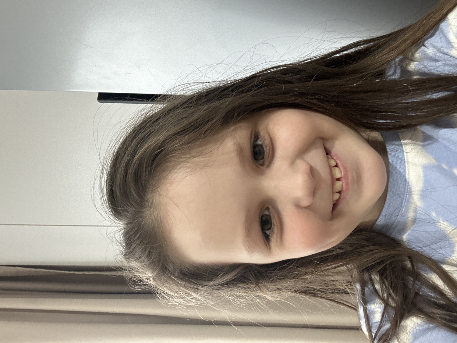 The image is a close-up portrait of a young girl, smiling broadly and looking directly at the camera. The photograph is oriented 90 degrees counter-clockwise, making the girl's head appear tilted. She has long, straight brown hair that frames her face and falls towards the right side of the frame. Her dark brown eyes are open, and her wide smile reveals both her upper and lower teeth, which suggest she is a child, possibly with developing permanent teeth.
The girl is wearing a light blue, long-sleeved garment with a white, patterned design, resembling a tie-dye or cloud pattern. Her expression is joyful and cheerful.
The background is softly blurred and indicates an indoor setting in Chaikovsky, Russia. To the upper left of the frame (when viewed in its current rotation), there is a light grey or white surface, possibly a wall or ceiling, with a subtle dark horizontal line, which might be an architectural feature or furniture edge. The lower left features a darker, textured beige or brown surface, potentially a curtain or another part of an interior wall. The lighting appears soft and even, consistent with indoor ambient light. No specific objects or text are visible apart from the girl and her attire.