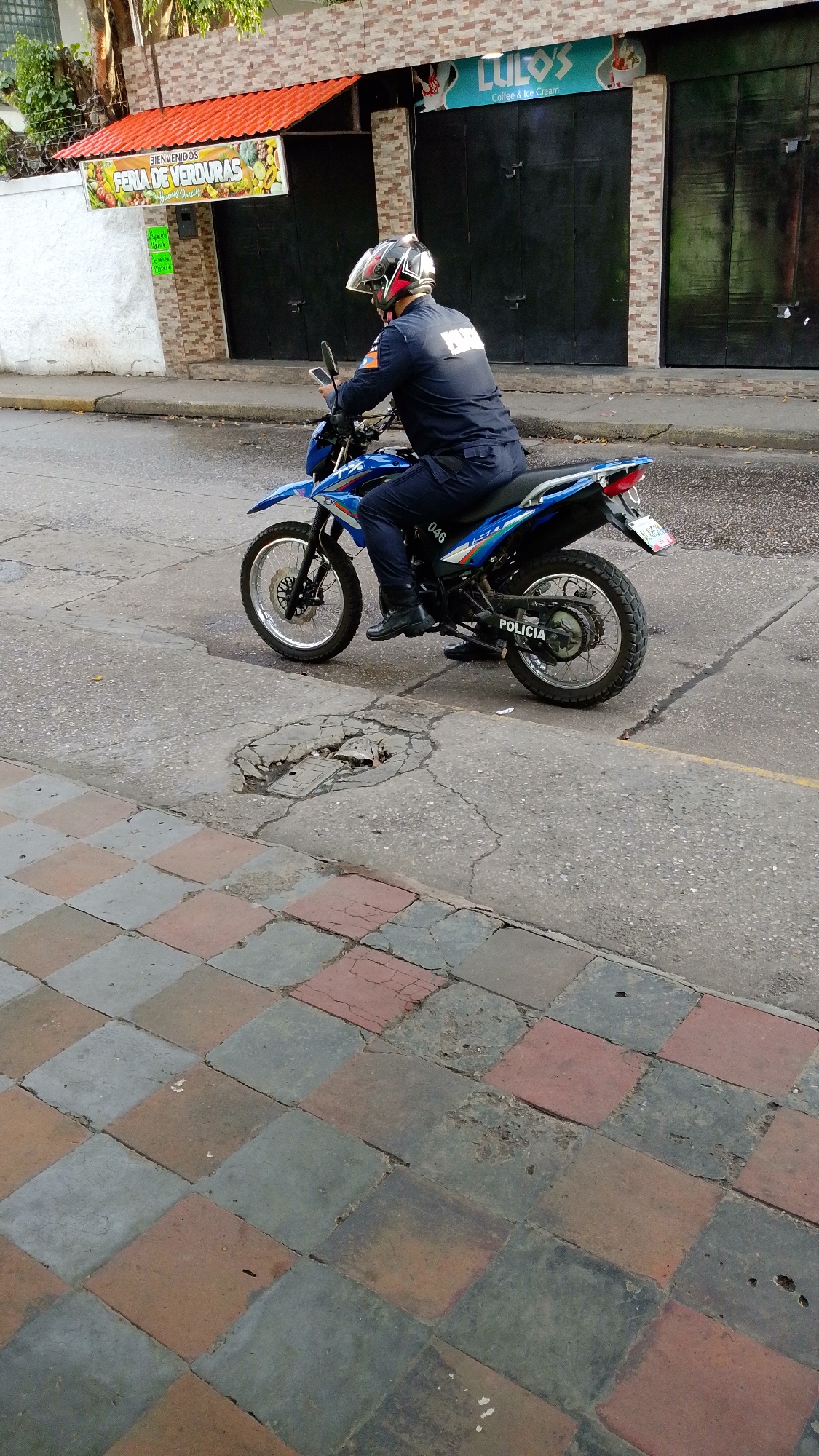 A police officer on a blue motorcycle is riding on a street in Tinaquillo, Venezuela. The officer is wearing a helmet and a dark uniform. The motorcycle has the word "POLICIA" (police) written on it, along with the number "046". The background shows buildings with storefronts, including a fruit and vegetable stand called "Feria de Verduras" and a cafe named "Lulos Coffee & Ice Cream." The street is paved, with a checkered pattern sidewalk in the foreground.