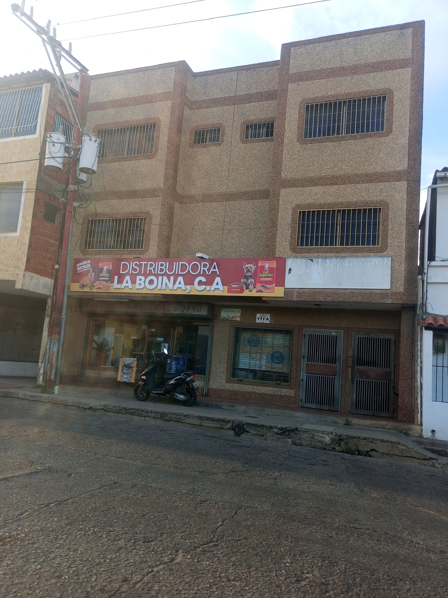 The image shows a three-story building in Juan Griego, Venezuela. The building has a tan, textured facade with brown trim around the windows and edges. The windows are barred. A sign above the storefront reads "DISTRIBUIDORA LA BOINA, C.A" and advertises dog food. Next to it, there is another sign that reads "ADMINISTRACION TRIBUTARIA 'Héctor Fleming'". A scooter is parked in front of the building, on the sidewalk. The street is asphalt and appears to be on a slight incline. A utility pole with transformers stands to the left of the building. Other buildings are visible on either side.