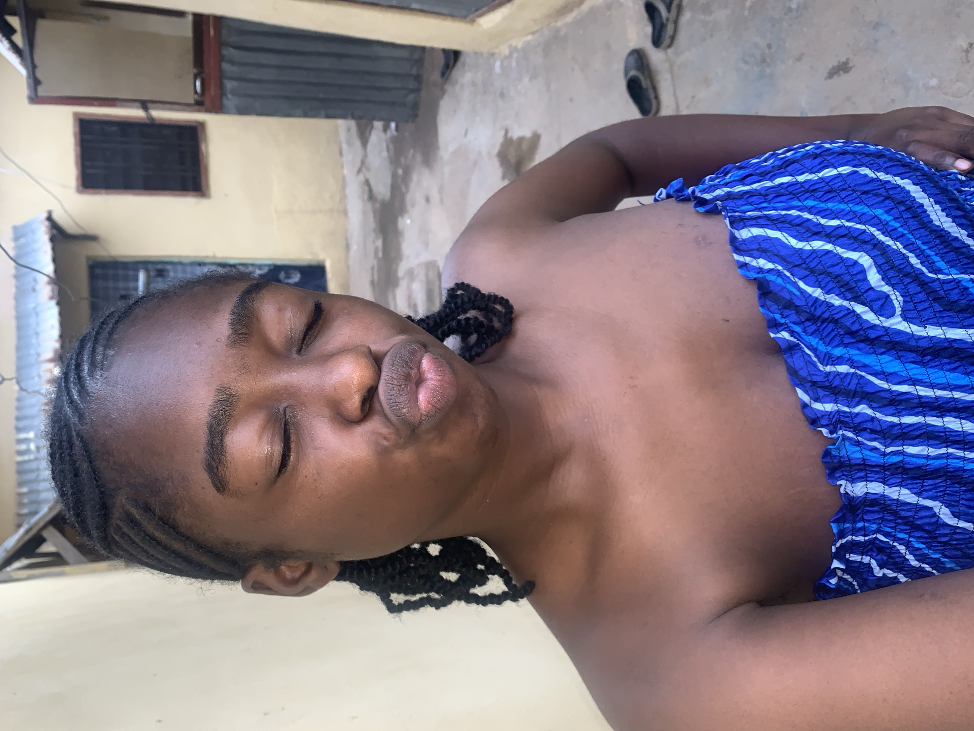 A young Black female is depicted in a close-up, waist-up selfie outdoors in Rafin Par, Nigeria. Her eyes are closed, and her lips are pursed forward in a pouting expression. Her hair is styled in dark cornrows on the scalp, transitioning to individual braids that rest on her shoulders. She wears a strapless, ruched top featuring a blue and white wavy striped pattern. The background consists of an unpaved outdoor pathway between structures. To the subject's left is a plain, light-colored wall. To her right and behind her are buildings with light-colored walls, dark-grilled windows, and sections of corrugated metal roofing. The ground is uneven and appears damp in areas. Multiple pairs of dark-colored slippers are visible on the ground in the right background. Faint lines, possibly indicating wiring, are present in the upper background against the wall and roof.