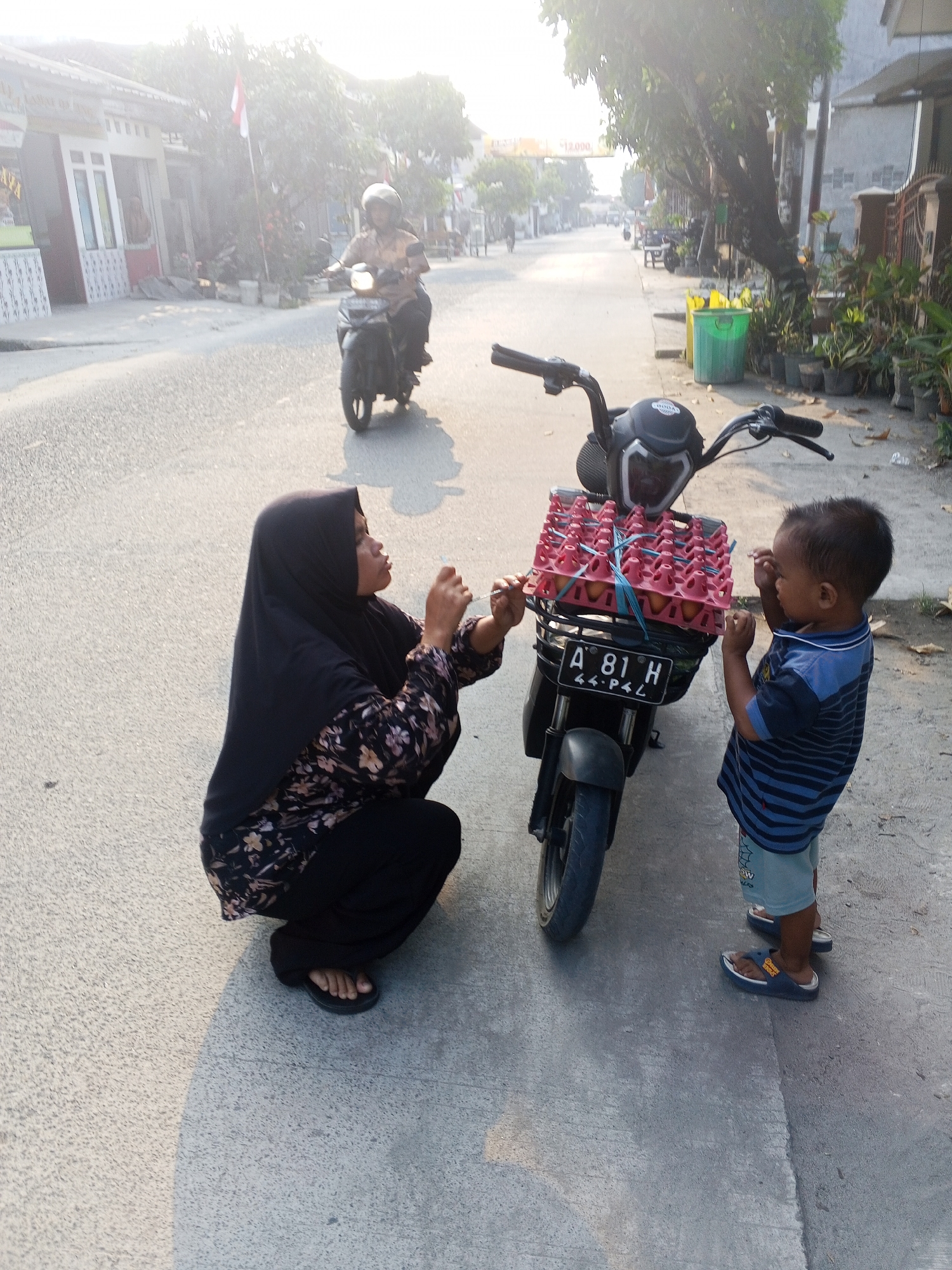 In a street scene in Ciruas, Indonesia, a woman wearing a black hijab and a floral patterned dress kneels on the pavement next to a black motorcycle. The motorcycle's front basket is filled with a pink egg carton containing eggs. A young child, wearing a blue striped t-shirt and light blue shorts, stands to the right of the motorcycle, facing the woman and interacting with her. In the background, a person rides a motorcycle away from the viewer, and houses and trees line the street. The scene is illuminated by the bright, hazy sunlight of what appears to be late afternoon.