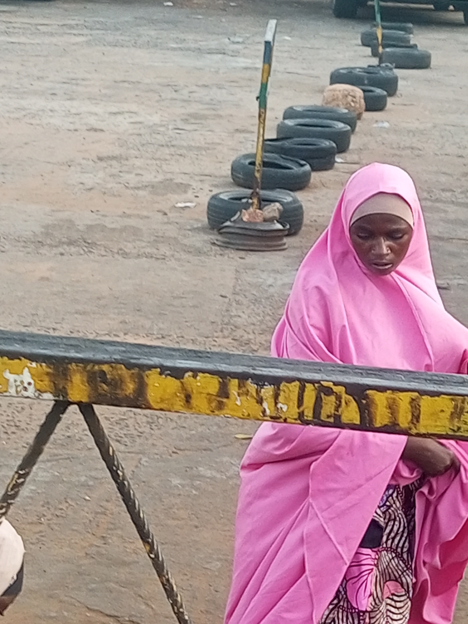 A woman wearing a pink hijab and a patterned dress stands next to a yellow and black metal barrier. Behind her, a row of tires with metal posts and stones is visible on a sandy ground. The location appears to be an outdoor area in Tempure, Nigeria.