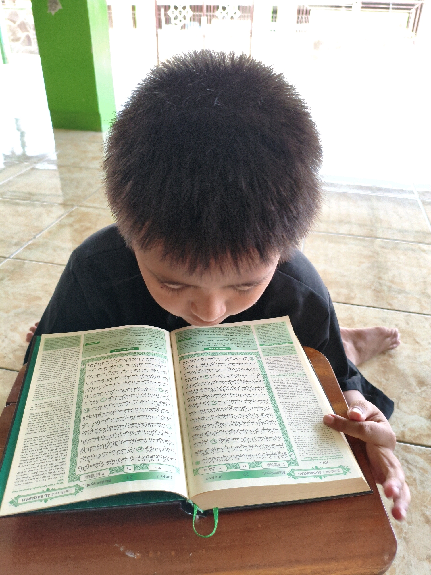 An over-the-shoulder, medium shot shows a young boy with short, dark hair sitting on a tiled floor and reading a Quran on a wooden stand. The boy is wearing a black shirt. The Quran is open to a page with Arabic text, translated into Indonesian on the side. The setting appears to be outdoors, possibly in a mosque or prayer area in Salatiga, Indonesia, with a green wall visible in the background. The lighting is bright, suggesting daytime.