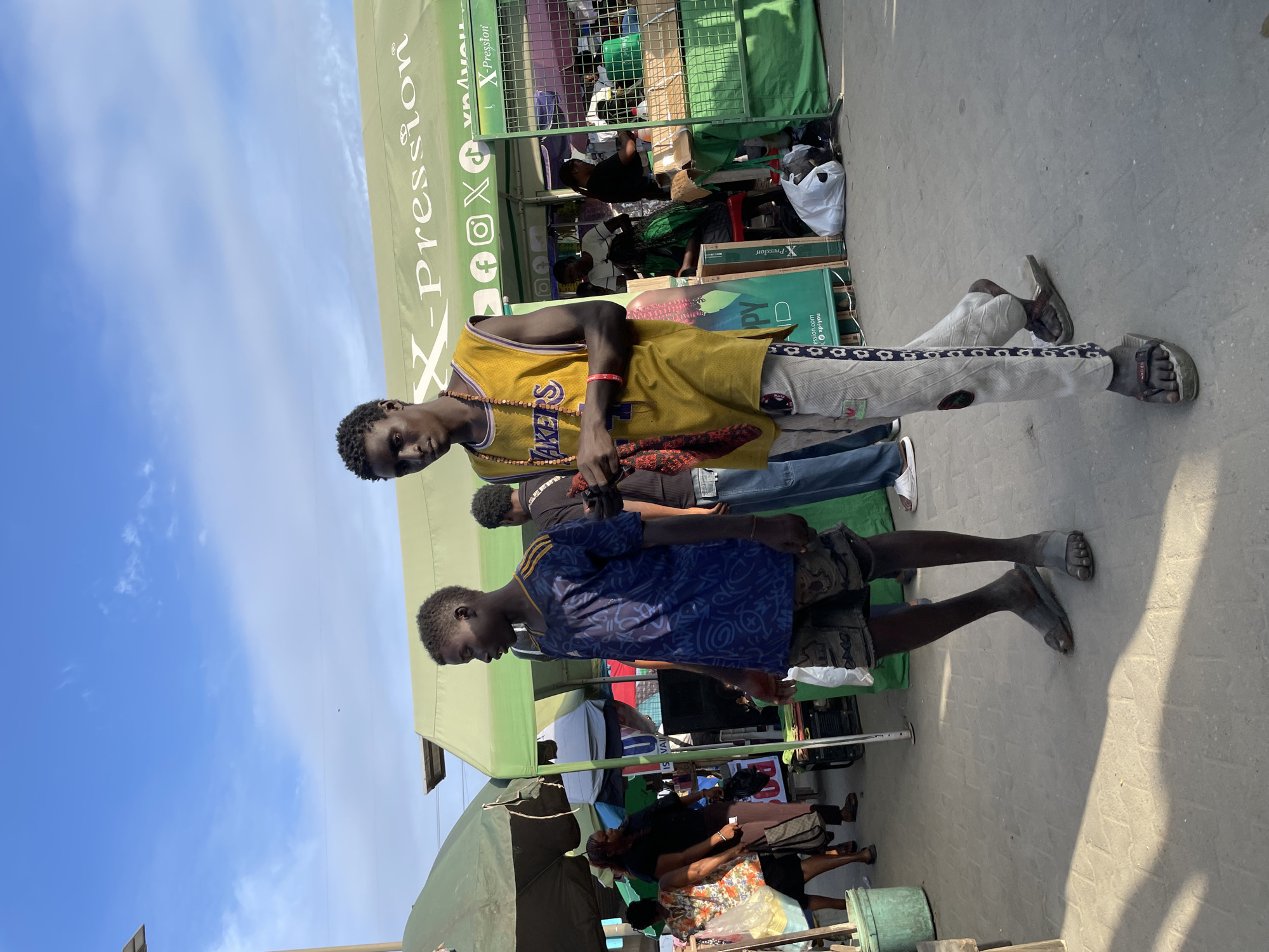Two young men are walking on a paved surface in what appears to be a market or street vendor area in Lagos, Nigeria. The man on the left is wearing a dark blue t-shirt with a repeating pattern and patterned shorts, along with dark sandals. The man on the right is wearing a yellow "Lakers" basketball jersey over a necklace, light-colored pants with a patterned stripe down the side, and dark sandals. He is holding a red cloth in his left hand. In the background, several market stalls with green awnings are visible. One awning has the text "X-Pression" on it, along with social media icons. Other people are visible in the background, some tending to stalls or walking. The sky is blue with some clouds. The lighting suggests it is daytime, possibly late afternoon, with strong shadows on the ground.