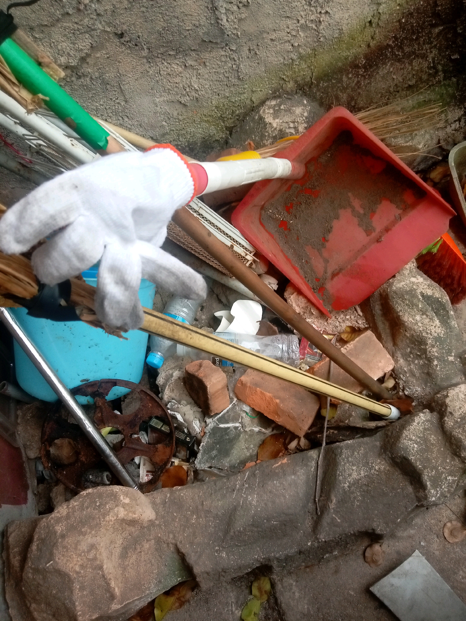 The image depicts a cluttered pile of cleaning supplies and debris near a rough concrete wall. A white glove, possibly a work glove, is prominently positioned in the foreground, obscuring part of a broom and a blue bucket. A red dustpan, filled with dirt, lies to the right, alongside a broom with orange bristles. Various construction materials, including bricks and large rocks, are scattered around. A metal pole and a rusty circular object are also visible. The scene suggests an outdoor area, likely a yard or alleyway, given the rough textures and the presence of natural elements like leaves. The location is specified as Batam, Indonesia, indicating a tropical or subtropical environment.