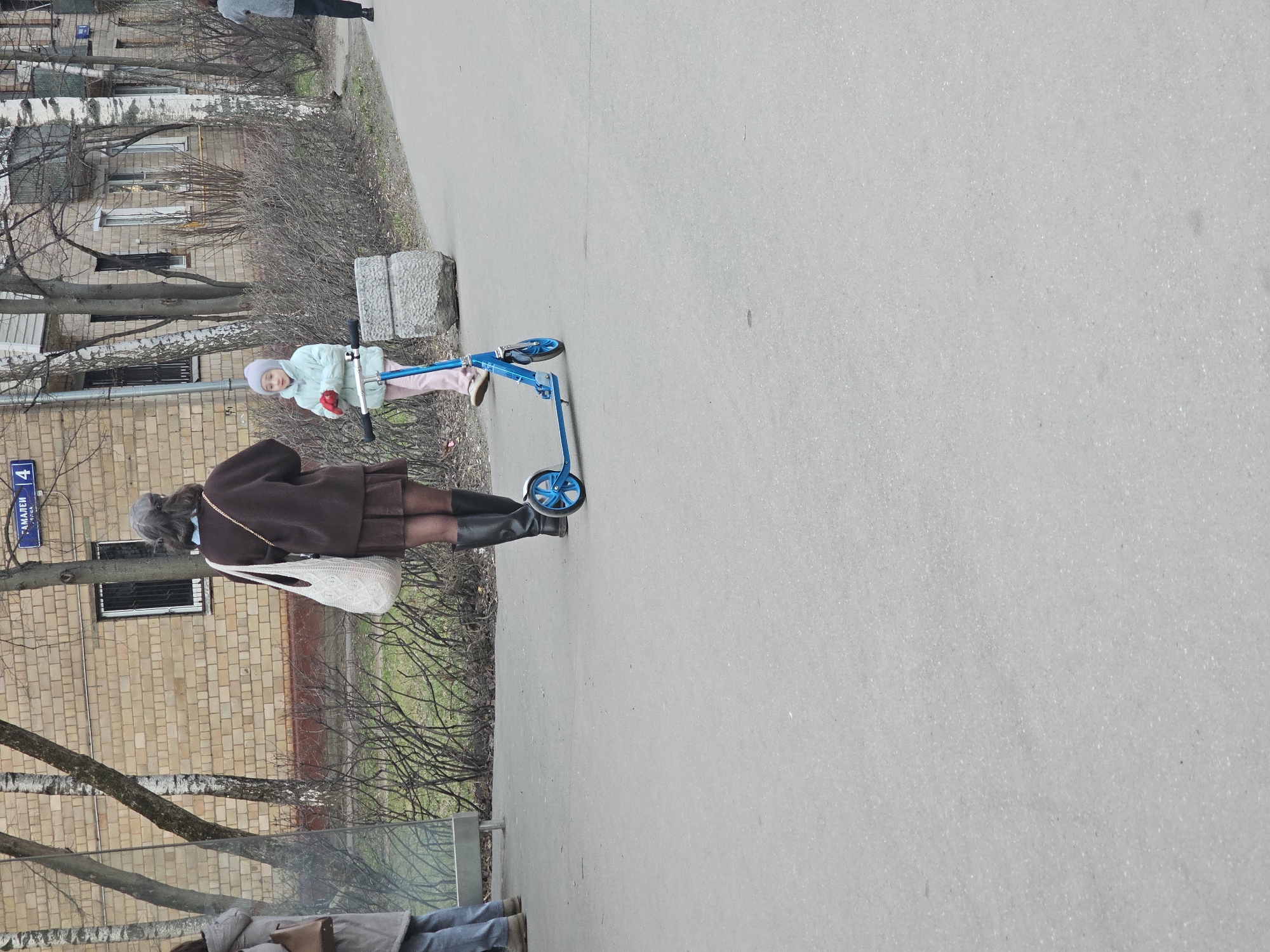 The image shows a girl on a blue scooter, next to an adult female. The woman wears a brown coat, black boots, and carries a white bag. Behind them is a brick building with bare trees. A street sign that reads "Амалеева улица, 4" ("Amaleeva street, 4") indicates the location is on Amaleeva street in Khimki, Russia. They appear to be on a sidewalk next to the building.