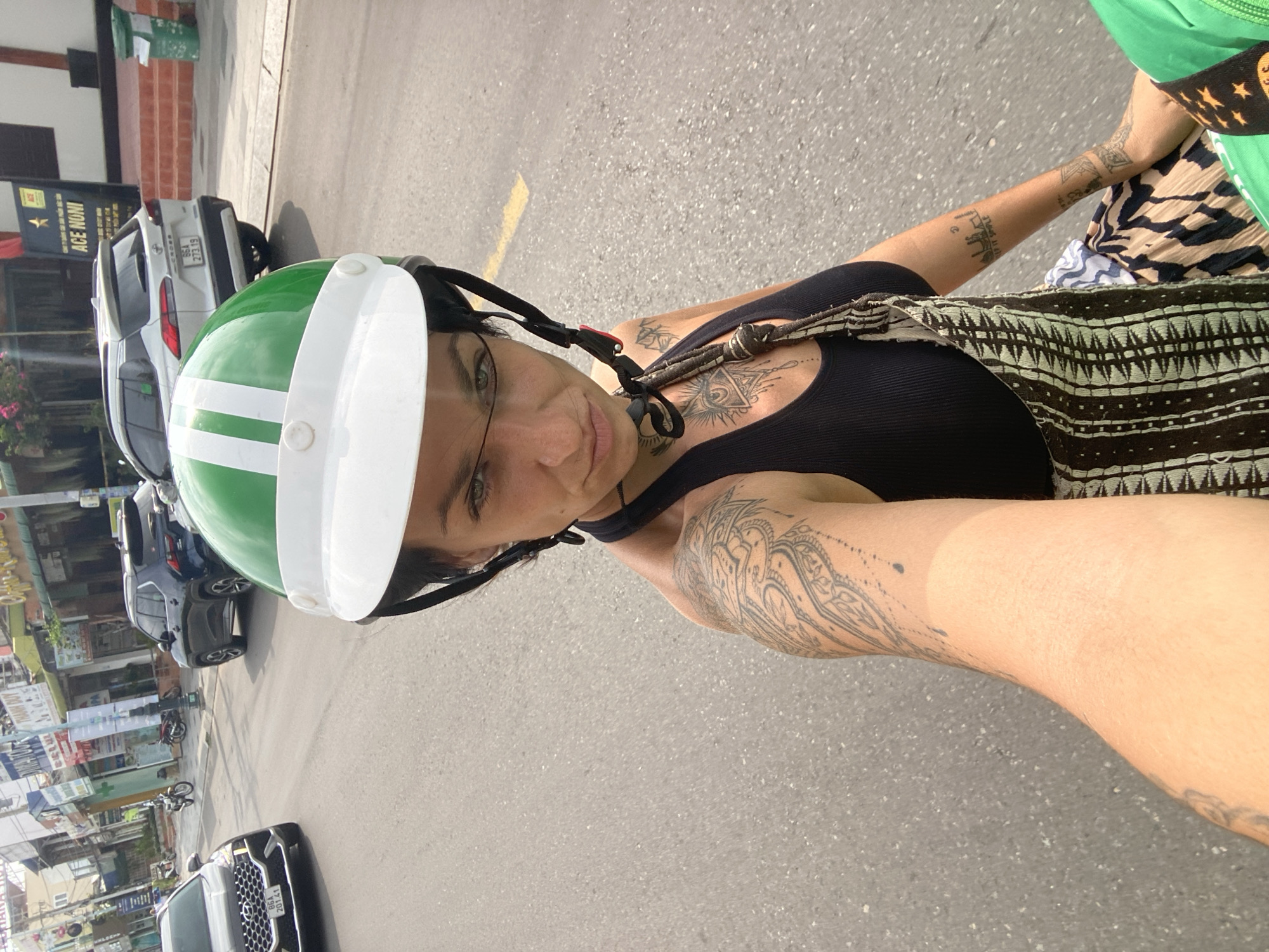 The image shows a woman with tattoos on her arm and chest wearing a green and white helmet and a black tank top with a patterned dress or apron. She is looking at the camera with a neutral expression. The background suggests a street scene in Phu Thuy Ward, Vietnam, with several cars, a motorcycle, and buildings visible. The road is paved and has yellow lane markings. The overall impression is a candid street-style photograph.