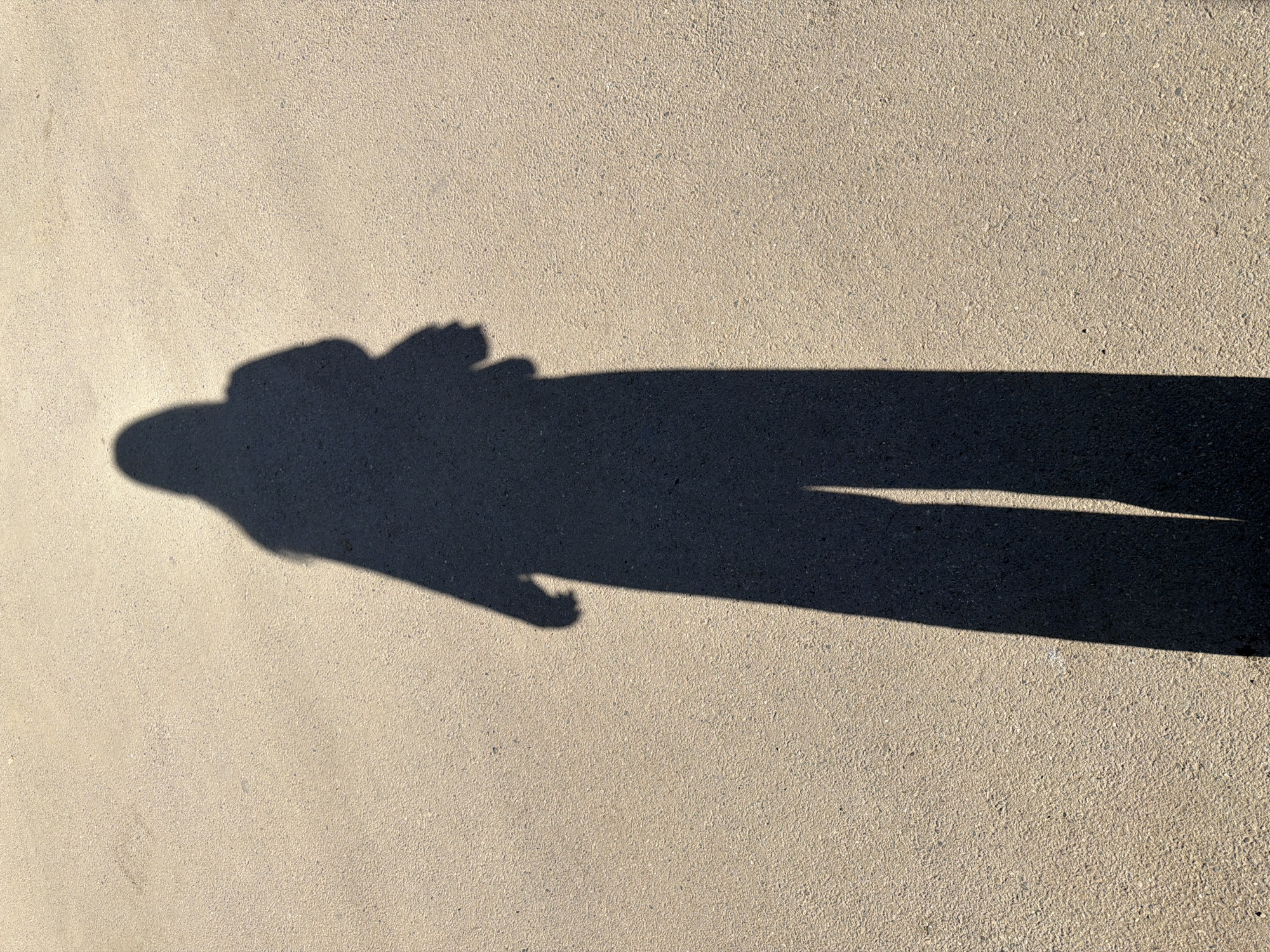 The image shows a shadow cast on a concrete surface. The shadow appears to be that of a person standing with their arms slightly raised or extended. The shadow is long and distinct, suggesting a low sun angle. The concrete is light-colored and textured. The location is unknown; no information suggests Krasnodar, Russia.
