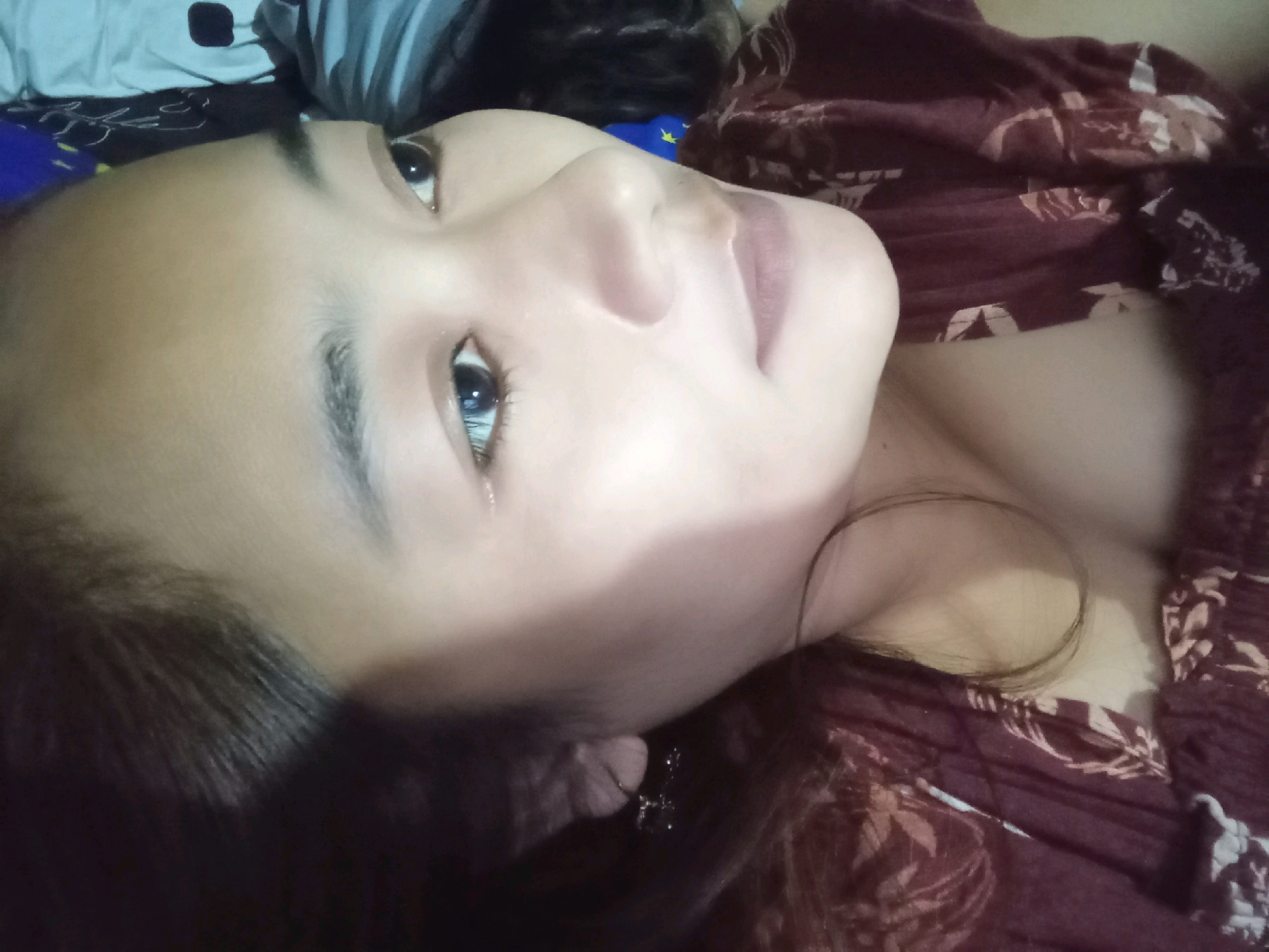 A close-up, low-angle shot shows a young person lying down, with their face angled towards the camera. They are on a bed with a patterned maroon comforter. The person has dark hair that is partially visible around their face and neck. Their eyes are open and looking towards the viewer. There is a hint of a necklace with a cross pendant resting on their chest. The lighting suggests it could be daytime or indoors. The overall mood is relaxed and intimate. The image was taken in Kedunglengkong, Indonesia.