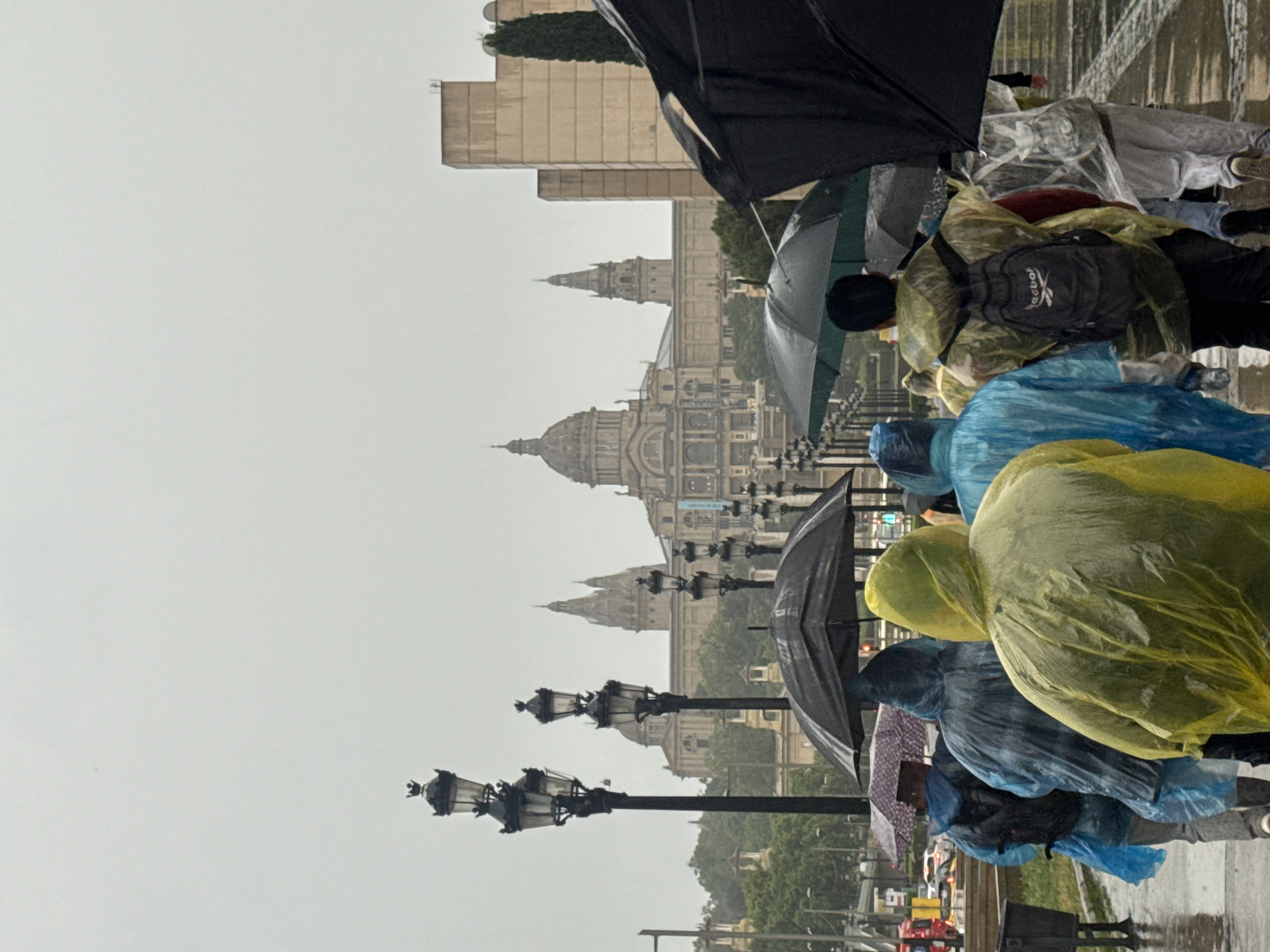 The image depicts a rainy day scene in Barcelona, Spain, with the Palau Nacional building visible in the background. In the foreground, a crowd of people wearing colorful rain ponchos and holding umbrellas are walking toward the camera. Streetlights line the path in front of the Palau Nacional, adding to the overall perspective of the scene. The sky is overcast, contributing to the rainy atmosphere.