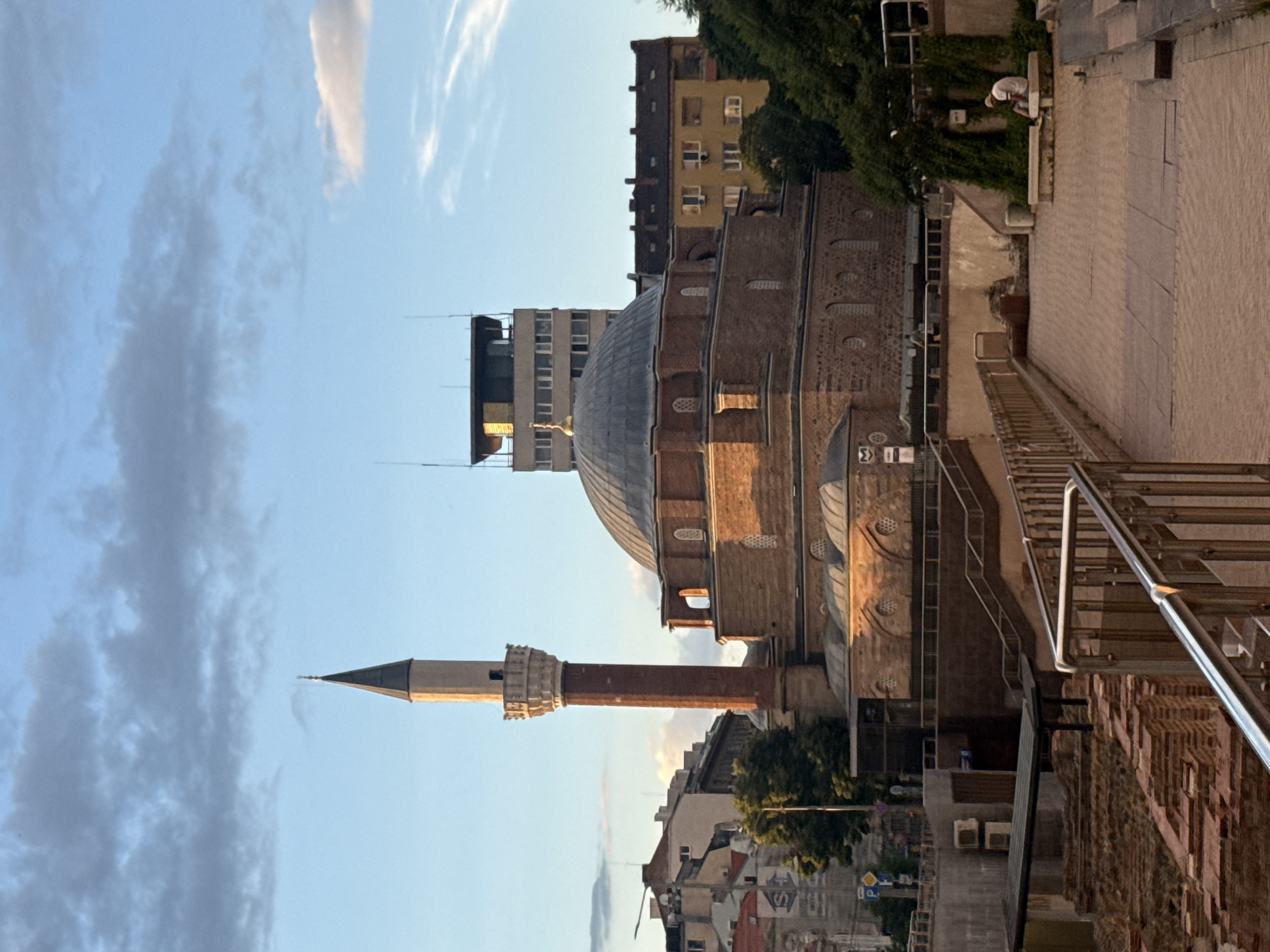 The image captures the Banya Bashi Mosque in Sofia, Bulgaria, under a blue sky with scattered clouds. The mosque, constructed of red brick, features a prominent dome and a tall, slender minaret. In the foreground, a paved walkway leads towards the mosque, bordered by metal railings and remnants of ancient brick structures. To the right of the walkway, a person is seated on a stone bench, appearing to rest. Behind the mosque, several multi-story buildings, including a taller, modern structure, are visible. The lighting suggests it is late afternoon or early evening, with the sun casting long shadows and illuminating the mosque's facade.