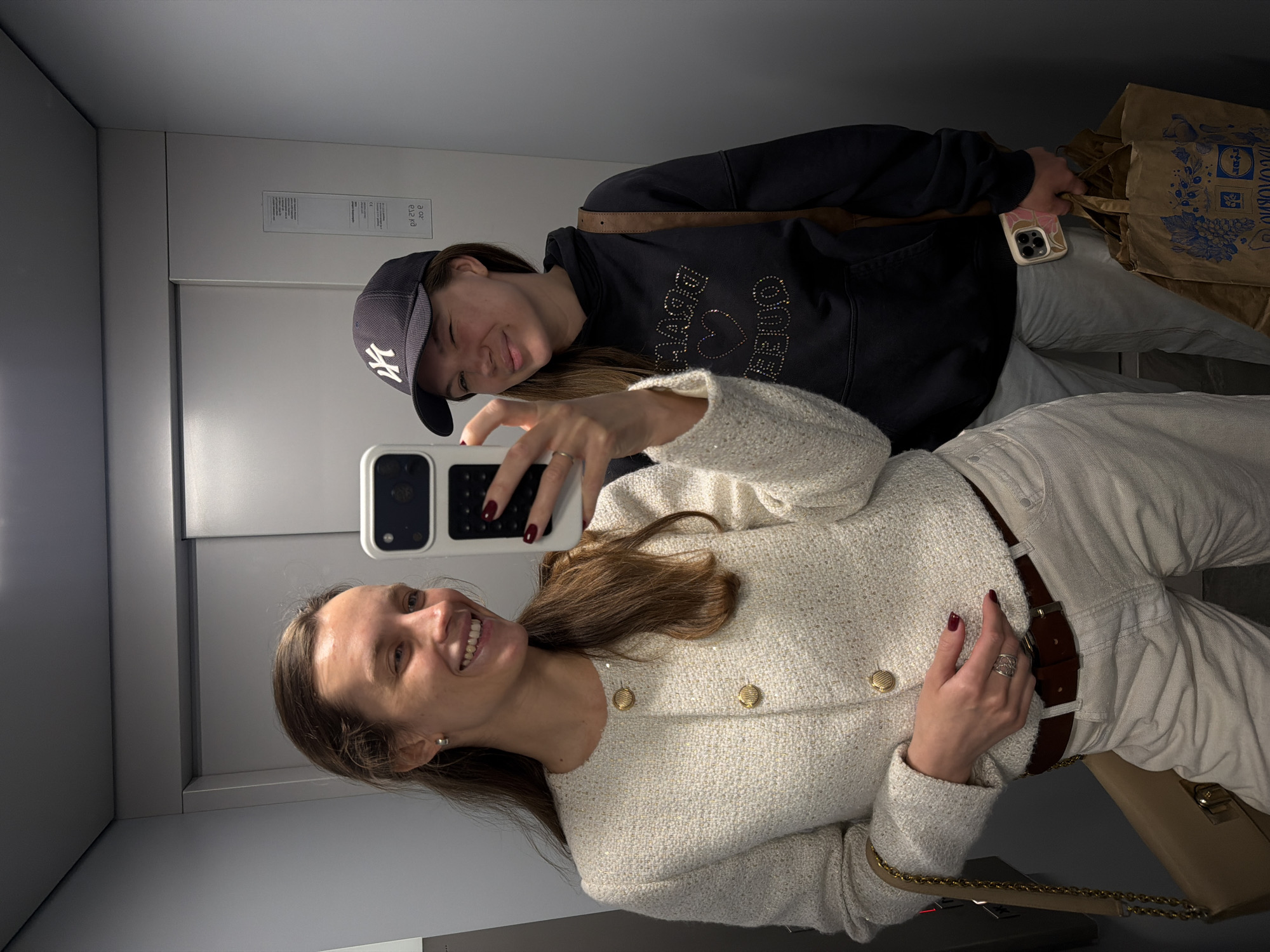 Two women are taking a selfie in an elevator. The woman in the foreground wears a white tweed jacket, a brown belt, and white pants. She is holding a phone with both hands. The woman in the background is wearing a black hoodie with a "Dreamer" inscription, a navy blue baseball cap with a white "NY" logo, and grey pants. She is also carrying brown paper bags. The elevator has grey walls and a white door. The location is an urban area of the city of Vozdovac Urban Municipality, Serbia.
