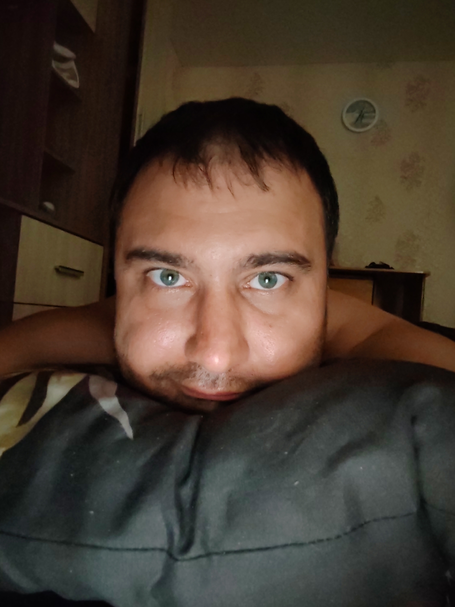 A close-up, head-on, front-facing, medium shot features a light-skinned man with short, dark hair and green eyes, lying on a dark pillow with a light-colored pattern. He is looking directly at the camera with his cheeks puffed out. The man's forehead, eyes, nose, and mouth are visible, with his chin resting on the pillow. The background is dimly lit, showing a wooden dresser to the left with drawers and shelves, and a wall with a floral wallpaper pattern and a circular clock with Roman numerals to the right. The scene is set indoors.