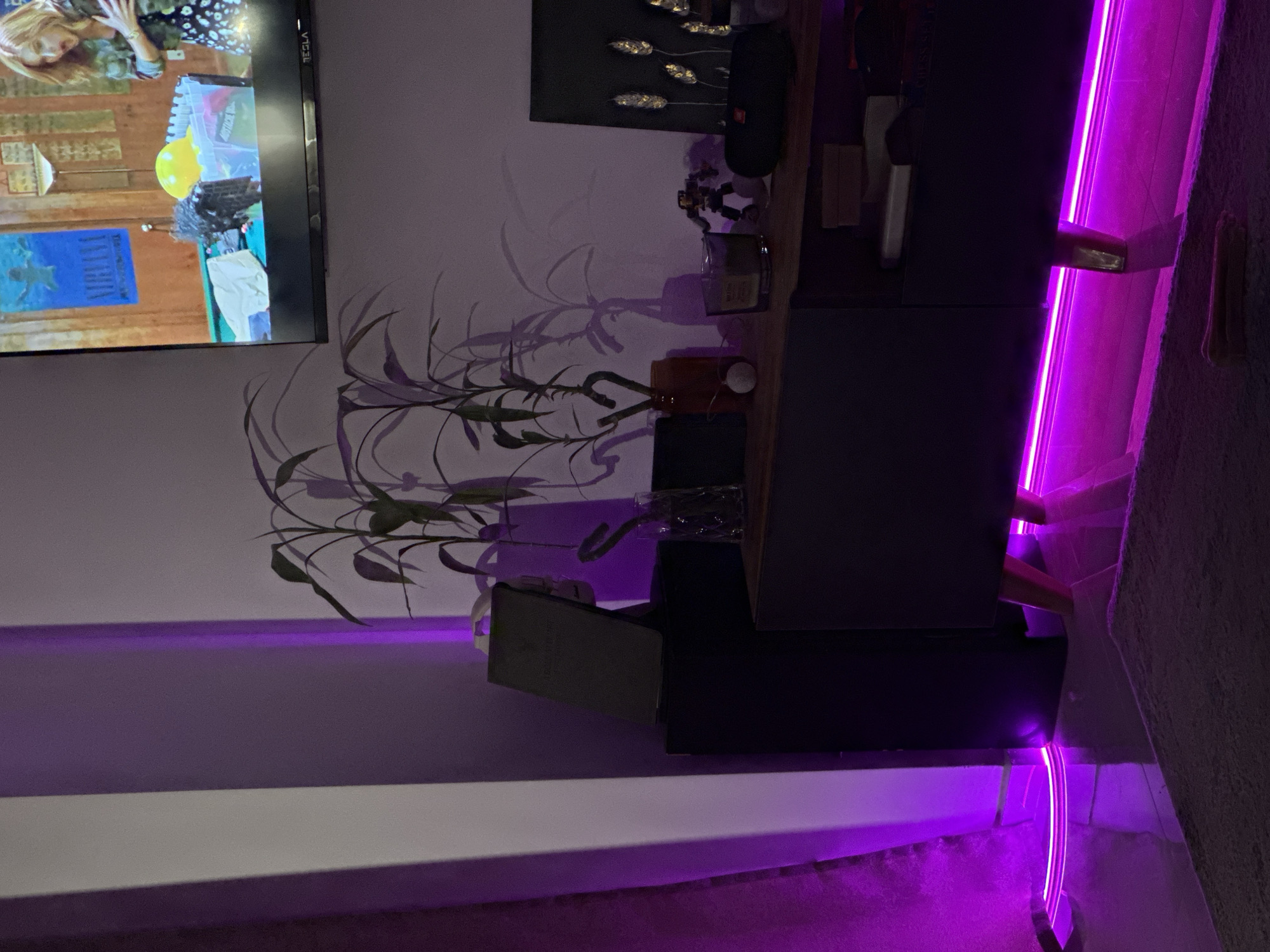 The image shows an interior scene at night, likely in a residence in an unknown location. A television screen displaying a scene from a show is mounted on the wall. In front of the television is a dark-colored entertainment center or shelving unit. On top of the unit are decorative items, including a plant in a vase, artwork, and other small objects. Purple LED strip lighting is visible behind and under the unit, casting a vibrant glow on the wall and the nearby rug. The room has a modern aesthetic with clean lines and minimal clutter. No people or indication of specific interaction are visible in the image.