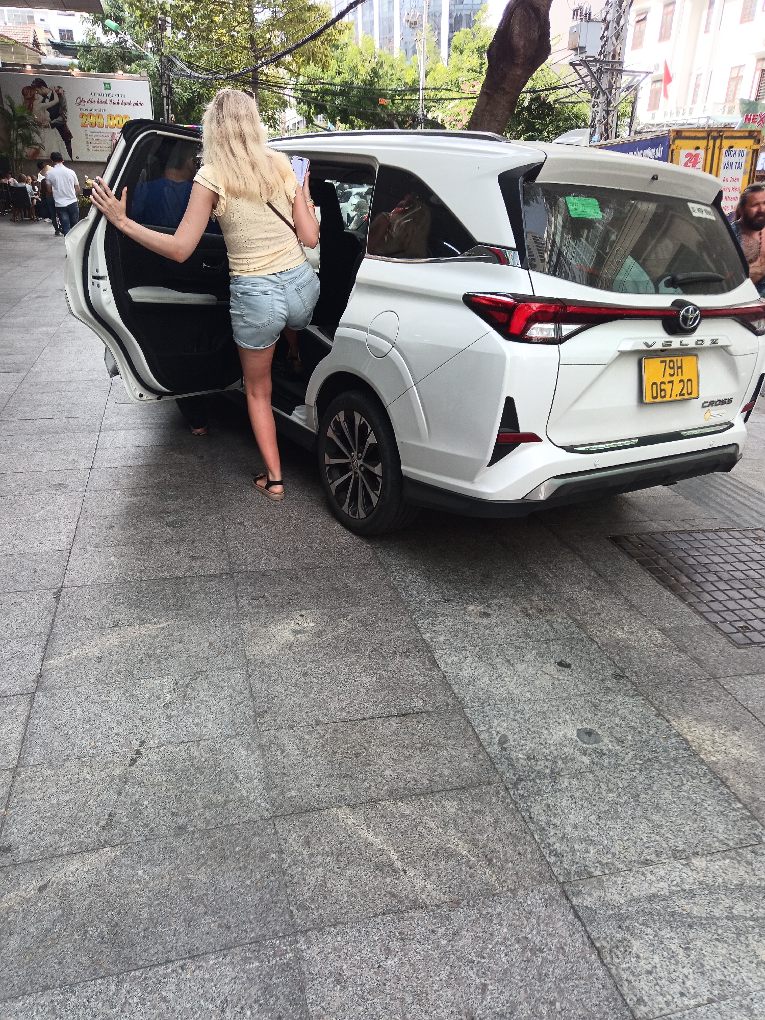 In Nha Trang, Vietnam, a blonde woman in a light yellow top and denim shorts is entering a white Toyota Veloz Cross. Her left hand is on the open driver's side door, and her right hand is holding a smartphone. The car is parked on a granite-tiled sidewalk adjacent to a street. The background shows a street scene with trees, buildings, power lines, and a large billboard. A man with a beard and no shirt is visible to the right of the car. The license plate of the car is 79H 067.20.