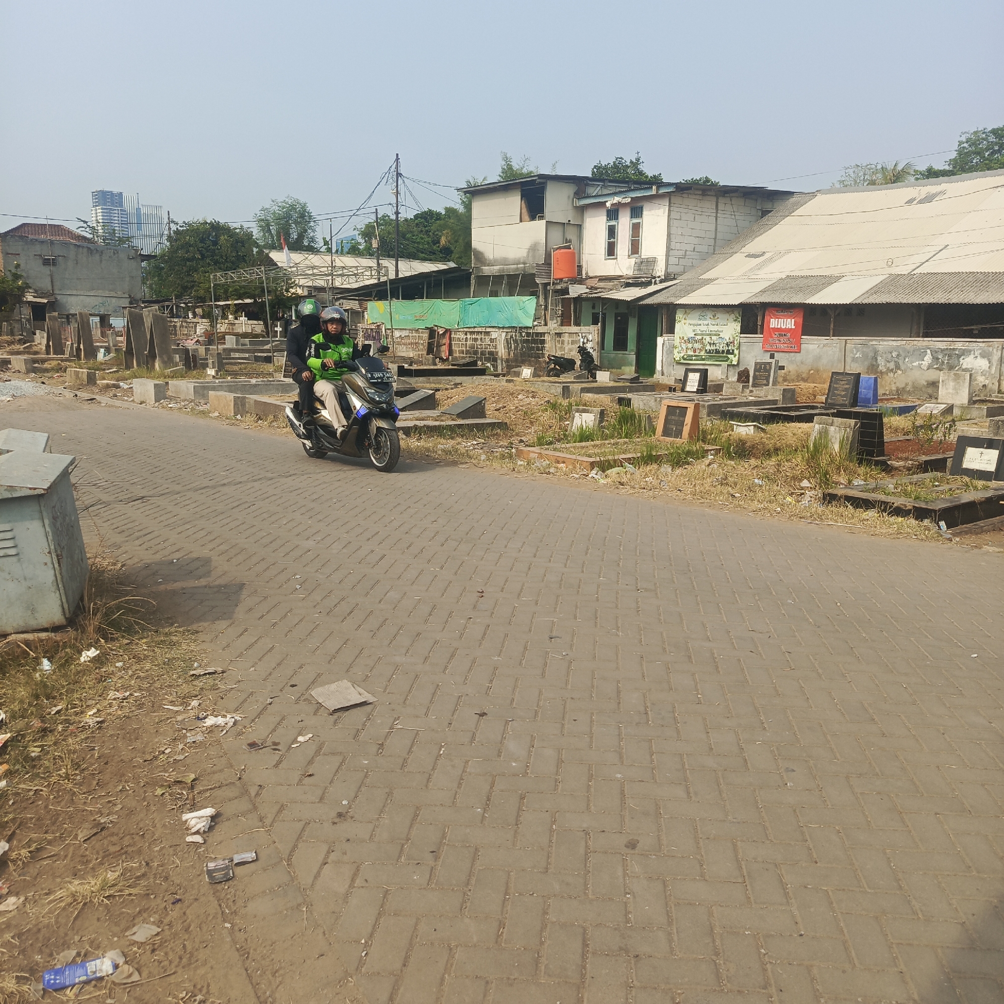 Two individuals are riding a scooter on a paved road adjacent to a cemetery. The person in front is driving, and the person behind is a passenger. Both are wearing helmets, with the driver wearing a green jacket with the Gojek logo visible. The cemetery contains numerous tombstones and grave markers, some of which are simple concrete slabs and others that are more elaborate. In the background, there are several buildings, including what appears to be a multi-story structure and some lower-lying buildings with corrugated metal roofs. Some trees are also visible. The road is made of interlocking concrete pavers, and there is some dry grass and scattered debris along the side of the road. The sky is clear and blue.
