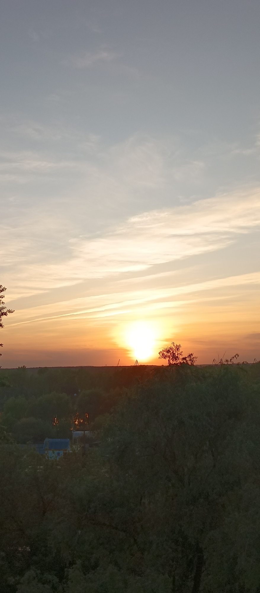 The image depicts a sunset scene over a landscape of trees and a building in the city of Unknown, Unknown. The sun is positioned on the horizon, emitting a bright, golden-orange glow. The sky above transitions from a light blue to an orange hue near the horizon. Below the horizon, there's a dense cluster of trees, and in the lower part of the image, there's a building partially obscured by the trees. The details of the building and the surrounding environment are not clearly visible due to the distance and the shadows cast by the trees.